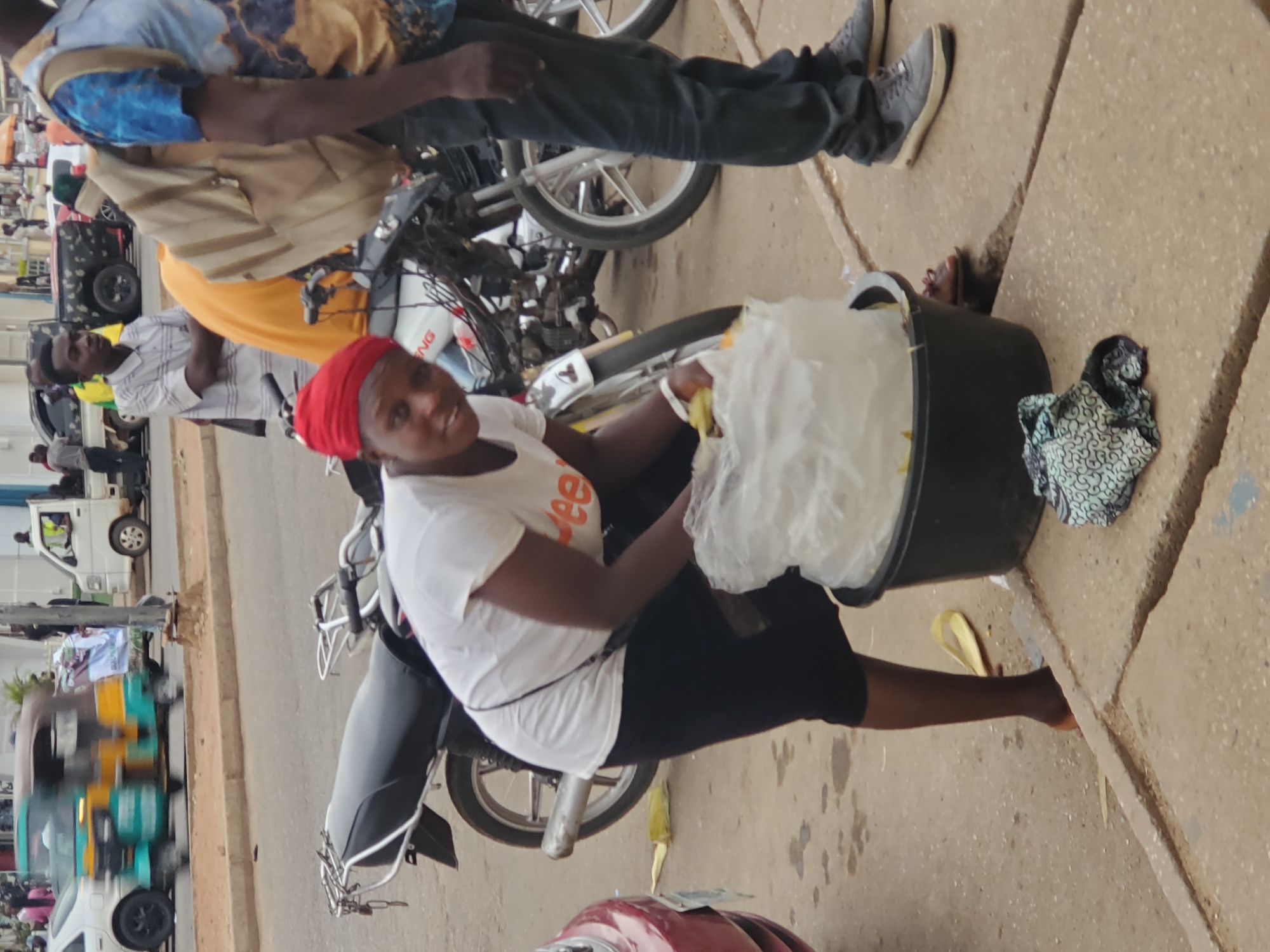 The image depicts a street scene, likely in Jos, Nigeria. A woman wearing a white t-shirt, black leggings, and a red headscarf is seated on a parked motorcycle. She is holding a large black bucket containing what appears to be food items wrapped in plastic. Beside the bucket on the ground is a patterned fabric. Other people are visible in the background, along with parked vehicles, including cars and motorcycles. The setting appears to be a busy street with a mix of pedestrian and vehicular traffic. The road is paved, and a sidewalk is visible on the right side of the frame.