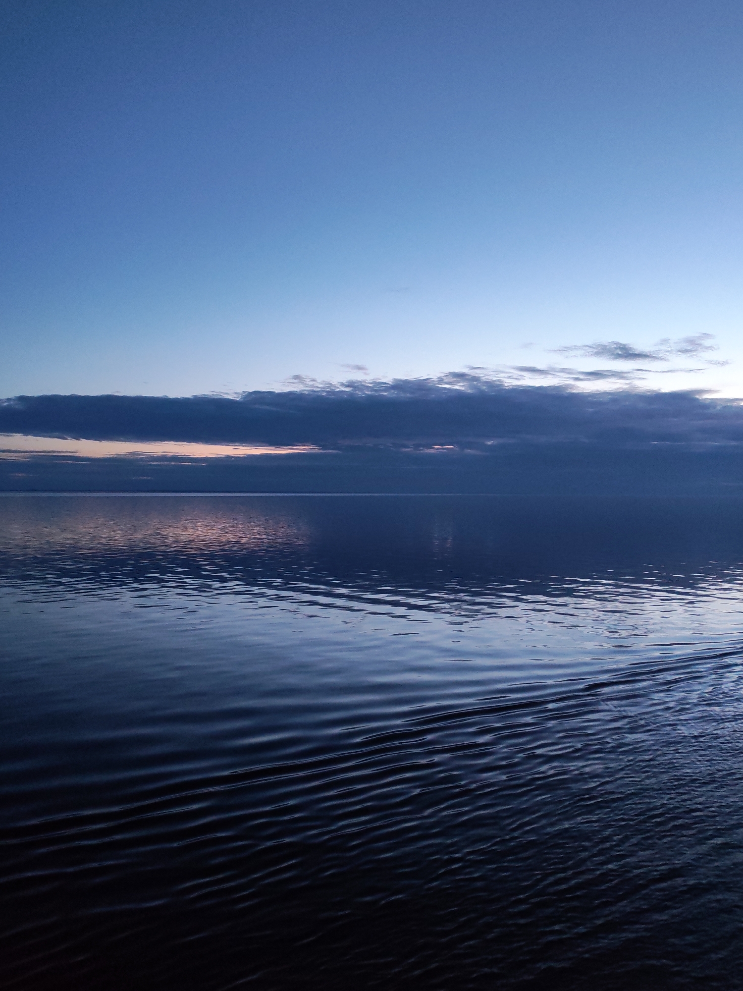 The image presents a tranquil twilight scene over a vast body of water, likely a large lake or sea, in an unknown location within Russia. The sky occupies the upper third of the frame, displaying a gradient from a deep, dark blue at the top to a lighter, almost pale blue near the horizon. A prominent, dark, elongated cloud band stretches horizontally across the sky, with glimpses of soft, lighter hues, possibly orange or pink, peeking through along the horizon. These colors suggest the quiet moments just after sunset or before sunrise.
The lower two-thirds of the photograph is dominated by the dark, reflective surface of the water. The water appears a deep, inky blue, almost black in the foreground, mirroring the muted tones of the sky and clouds. Faint, ethereal reflections of the overhead clouds and the subtle light at the horizon are visible on the water's surface. In the foreground, particularly extending towards the right, distinct, parallel ripples or a wake disturb the otherwise calm water, hinting at recent movement, possibly from a boat that is not visible within the frame.
There are no people or specific objects discernible in the scene. The overall mood is one of profound peacefulness and expansive solitude. The weather appears calm, with a mix of clear sky and cloud cover. There is no visible text in the media file.