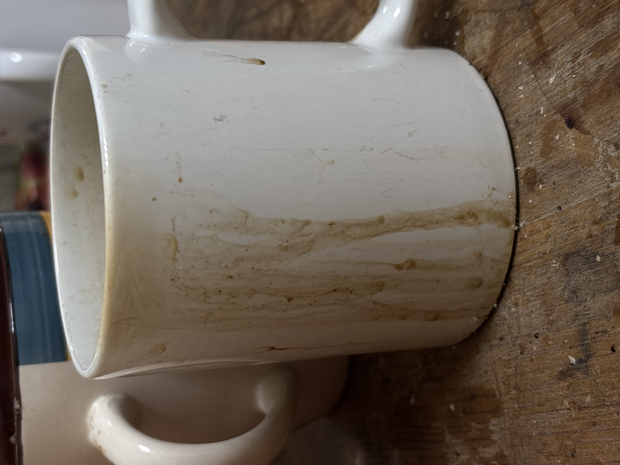 The scene depicts two ceramic mugs on a textured wooden surface, situated in what is identified as the city of Karasi, Russia. A white ceramic mug occupies the foreground, exhibiting extensive dried, dark brown liquid streaks and splatters vertically along its exterior from the rim to the base. A white handle is visible on the right side of this mug. Partially obscured behind and to the left of the foreground mug is a larger, light-colored ceramic mug, featuring a horizontal band of blue and brown coloration near its upper rim and a prominent light-colored handle on its left. Both objects rest on a wooden surface characterized by visible wear, dust, and scattered light-colored particulate matter. Near the base of the foreground mug, on the wooden surface, several small, dark, spherical objects are observable. The background elements are out of focus. No people are present.