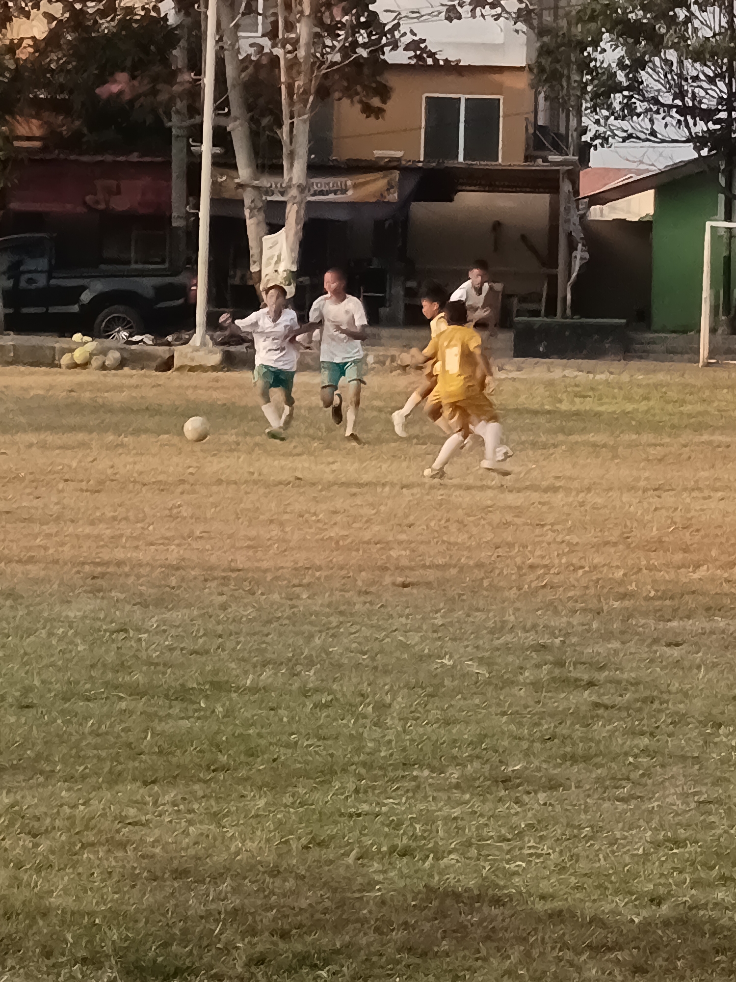 The image depicts a group of boys playing soccer on a grassy field in Desa Gunungsimping, Indonesia. In the foreground, a soccer ball rests on the dry, brown grass. Several boys are in motion, actively engaged in the game. Two boys in white shirts and green shorts are running towards the left of the frame, while another boy in a yellow shirt and white shorts is positioned towards the right, appearing to be dribbling the ball. In the background, trees line the edge of the field, and buildings and a dark-colored pickup truck are visible behind them. A white goalpost stands on the right side of the image. The lighting suggests it is late afternoon or early evening.