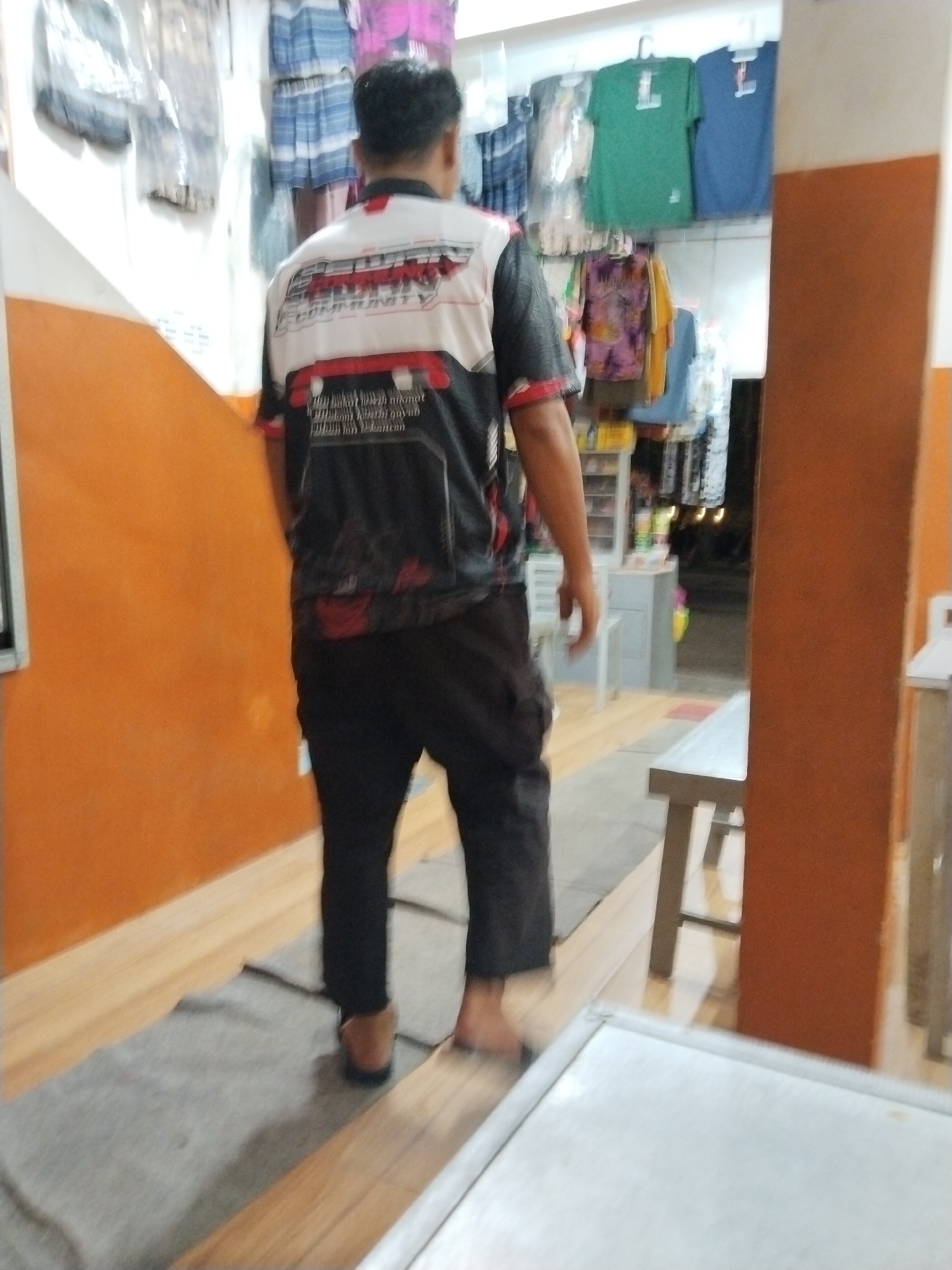 A man with dark hair and wearing a black shirt with red and white accents and black pants walks away from the camera. The shirt has writing on the back. He is indoors in what appears to be a small shop or market. There are clothes hanging on racks behind him, including green and blue shirts. The walls are orange. The floor is tiled with a grey mat and wooden planks. A wooden bench is visible to the right of the man. The scene is located in the city of Prigi, Indonesia.