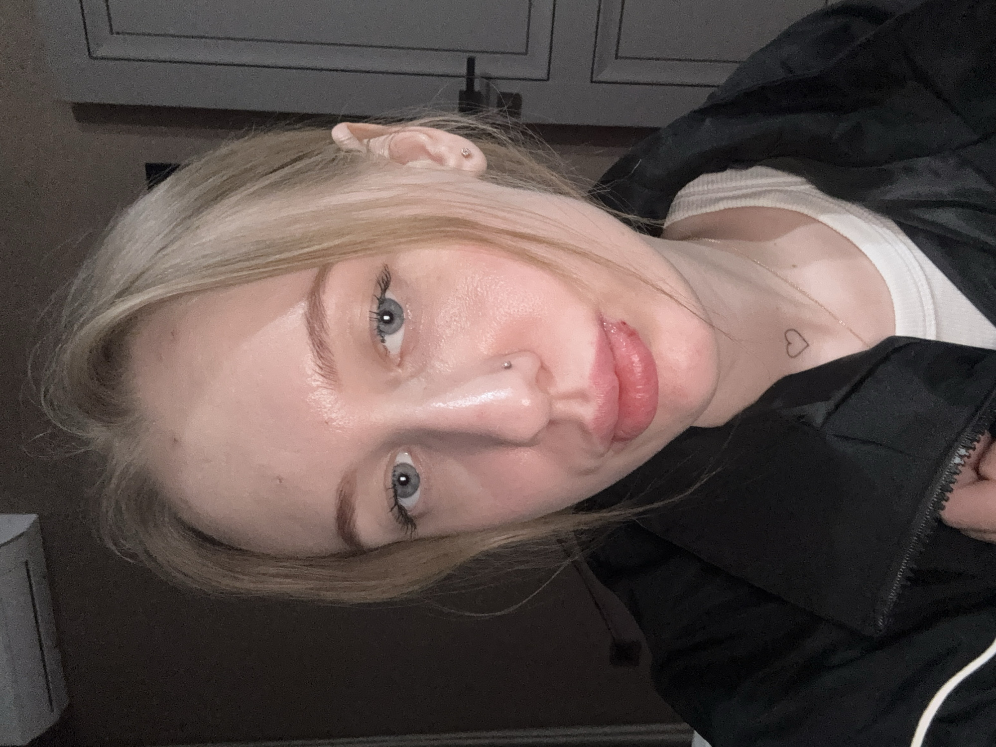 A close-up image depicts a young adult female indoors. Her blonde hair is parted on the right and falls partially over her forehead. She has blue eyes and fair skin. A small silver stud piercing is present in her right nostril, and a small stud earring is visible in her left earlobe. Her lips appear full, with a visible lesion or scab on the lower lip, slightly right of center. A small, outline heart tattoo is located on her neck, to the left of her midline. She is wearing a white top beneath an open black zip-up jacket, the zipper pull of which is held by her right hand. The background is an interior space, out of focus, showing a dark wall on the left with a portion of a light-colored, wall-mounted unit in the upper left corner. On the right, a light-colored door with a dark handle is visible. The exact location within the city of Azov, Russia, cannot be determined from the visual evidence provided.