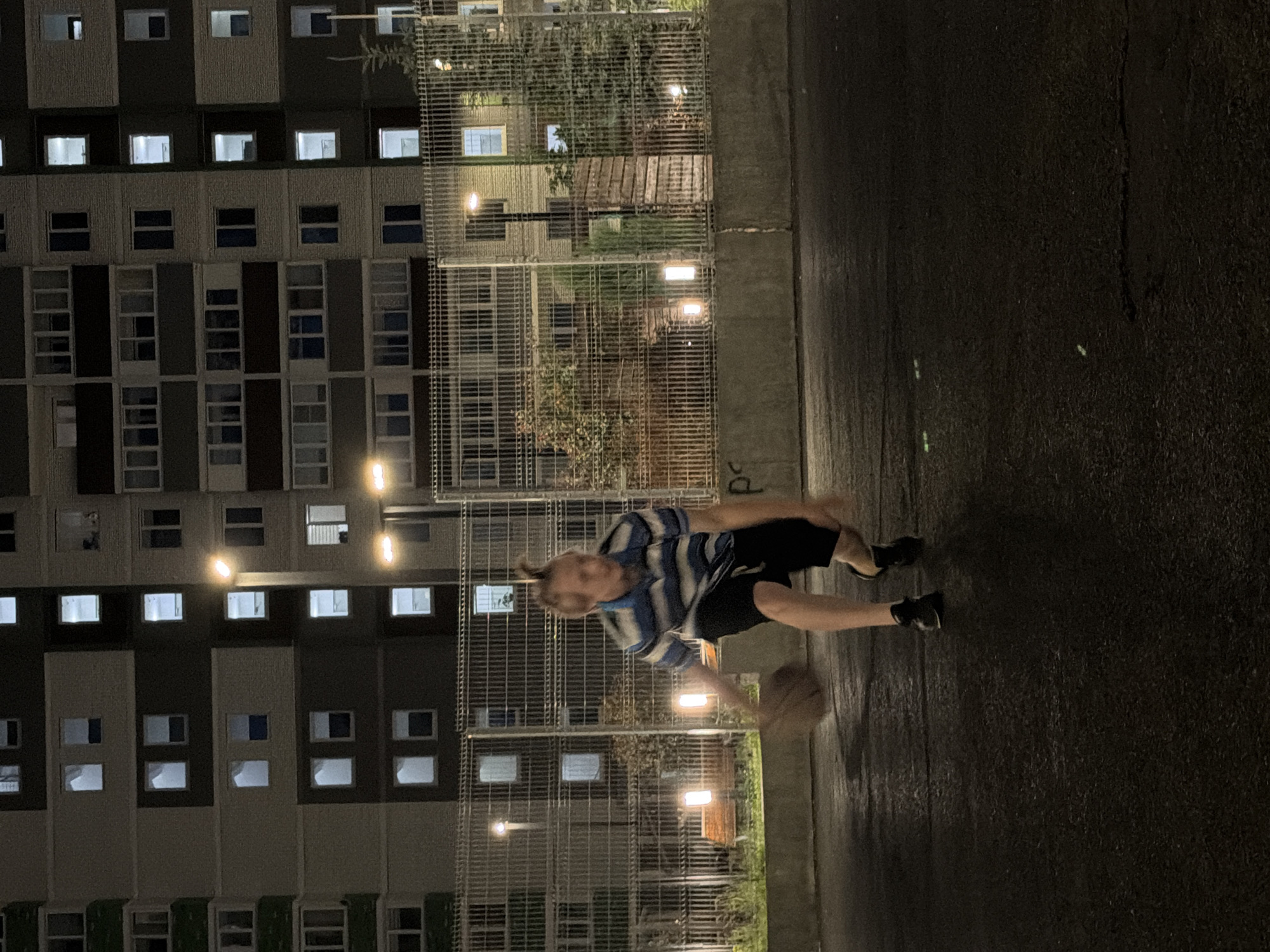 The image depicts a young man playing basketball on an outdoor court at night. He is wearing a blue and white striped shirt and black shorts. The court appears to be asphalt and is wet, reflecting the lights. Behind the court is a chain-link fence, and beyond that, a multi-story apartment building with many illuminated windows. There are also streetlights illuminating the court. The scene appears to be in an urban residential area in Izhevsk, Russia, given the architectural style of the building and the general urban setting. The man is in motion, with the basketball in front of him, suggesting he is dribbling or about to shoot. The overall atmosphere is one of casual recreation in an urban environment at night.