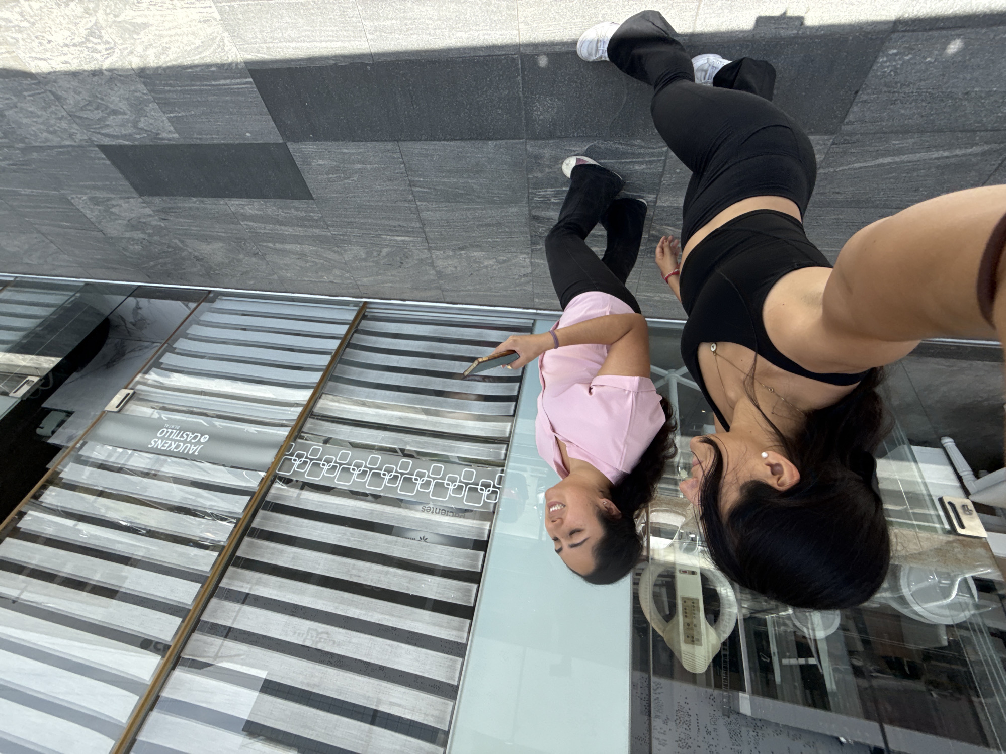 Two women are standing on a mirrored surface outside of what appears to be a store in Puebla, Mexico. The surface is reflecting their images and the surrounding environment. One woman is wearing a pink shirt and holding a phone; the other is wearing a black crop top and black pants. The store has multiple packages of blinds on display. Taukens Castillo Puebla is written on the blinds in the window. The ground is made of grey tiles.