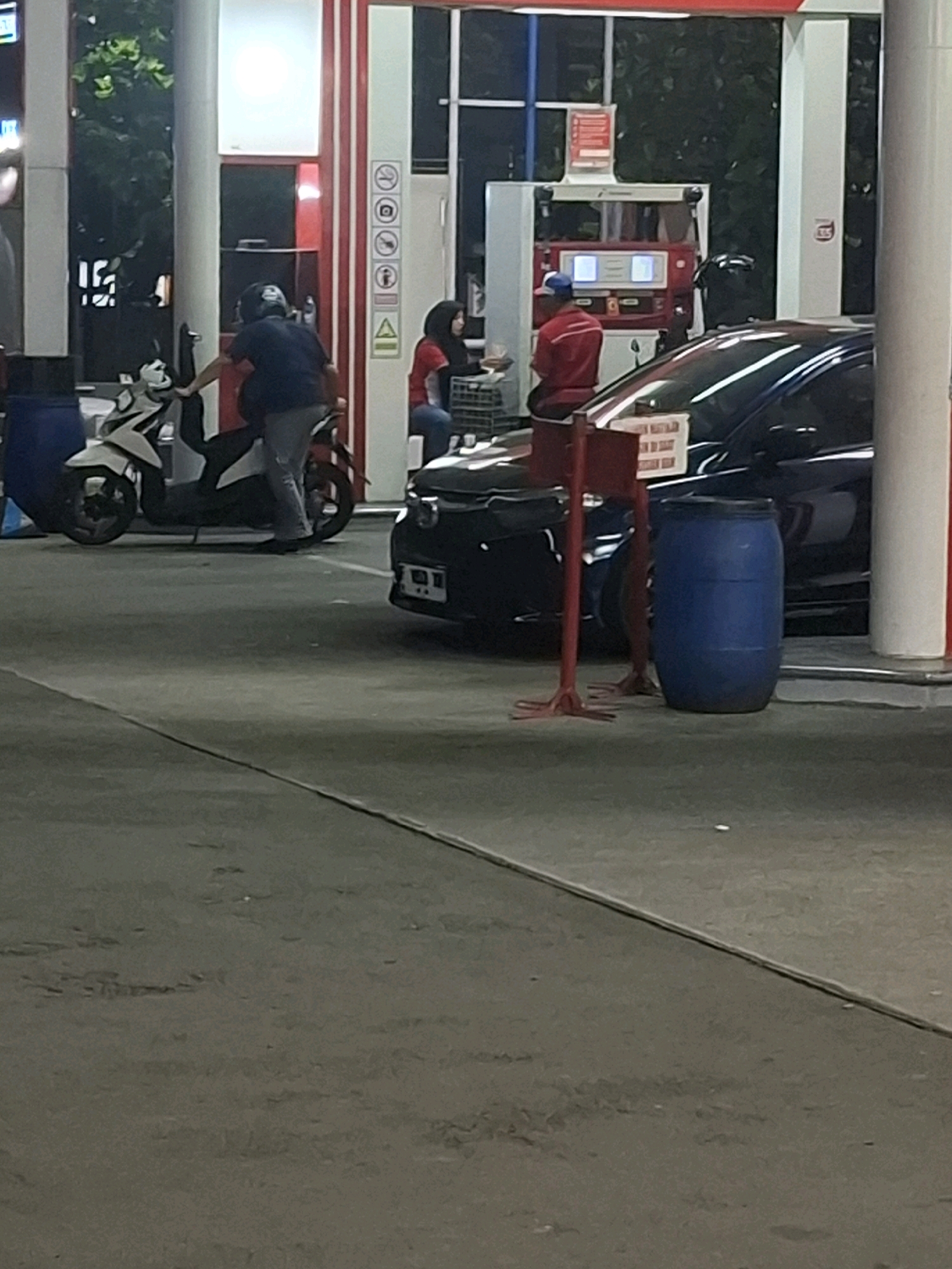 This is a medium-shot, outdoor, eye-level photograph taken at night, likely at a gas station in the city of Karangtengah, Indonesia. The scene depicts a dimly lit area with a fuel pump and several vehicles.
In the foreground, a black car is parked to the right. To the left of the car, a large blue barrel is visible, and a red metal stand with a white sign is positioned in front of the car.
In the midground, a white scooter is parked to the left, with a person wearing a dark blue shirt and a helmet leaning towards it. Behind the scooter, a person wearing a red shirt and a blue cap is seated. To the right of this person, a woman in a red shirt and headscarf is also visible, seated.
The background shows a section of a gas station building with white and red vertical columns and a brightly lit canopy overhead. Trees and foliage are visible behind the building. The ground surface appears to be asphalt.