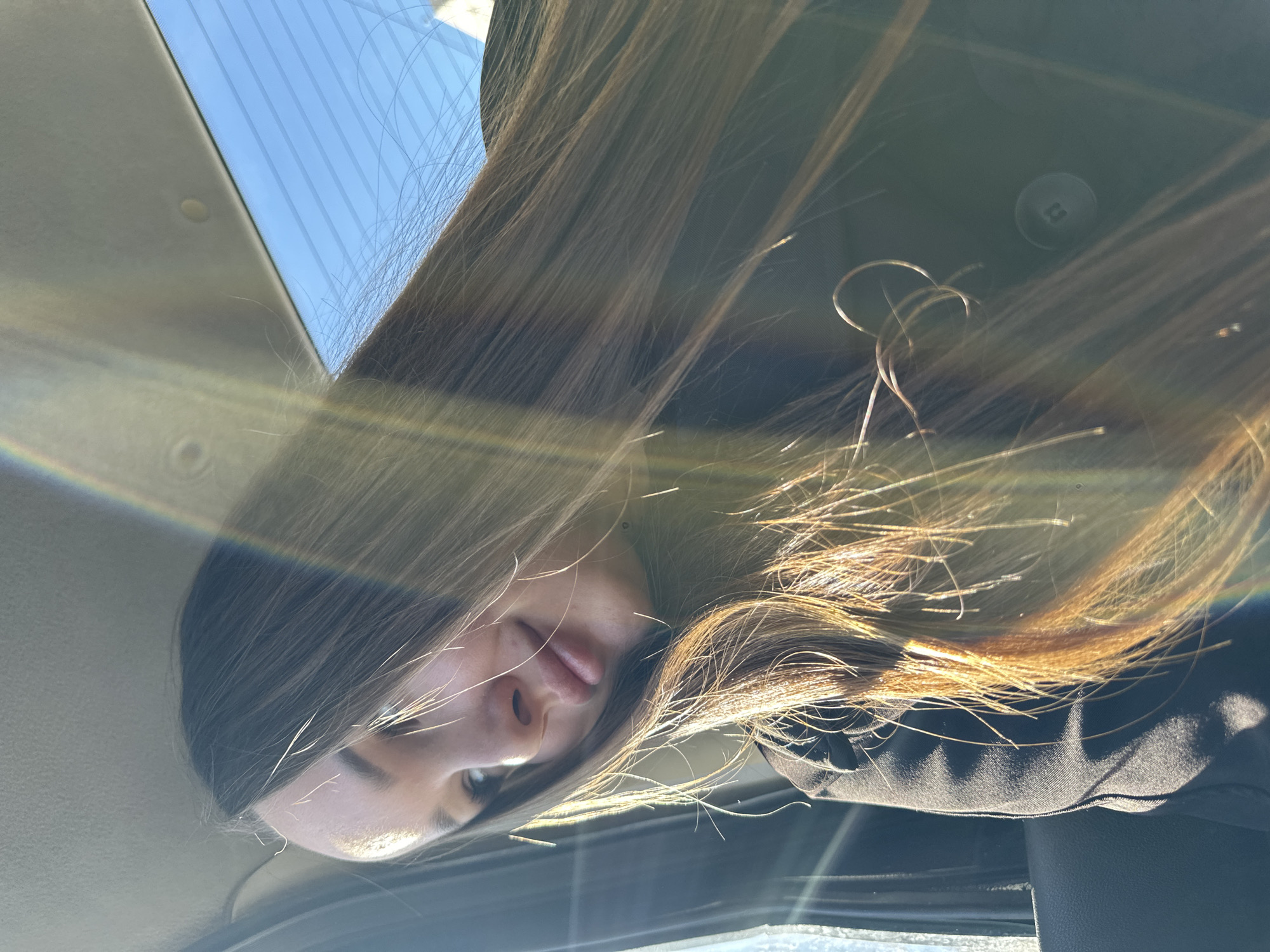 The image shows a woman with long brown hair, possibly inside a car. The scene is brightly lit with sunlight, creating a lens flare effect. The interior of the car is visible, including the roof and a button on what appears to be a jacket. The woman is partially visible, showing her face and hair. The location cannot be identified as Chaikovsky, Russia, based on the visual information provided.