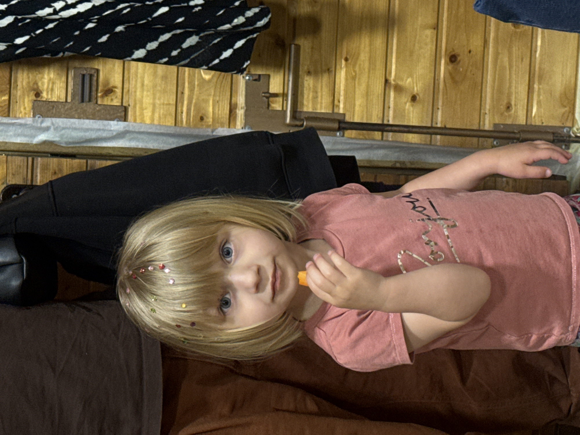 A young child with blonde hair, adorned with colorful sequins, is lying on a brown surface. The child is holding a small orange object. The background includes a wooden wall with attached metal fixtures, a black and white patterned item, and a black garment. The location is Unknown, Unknown.