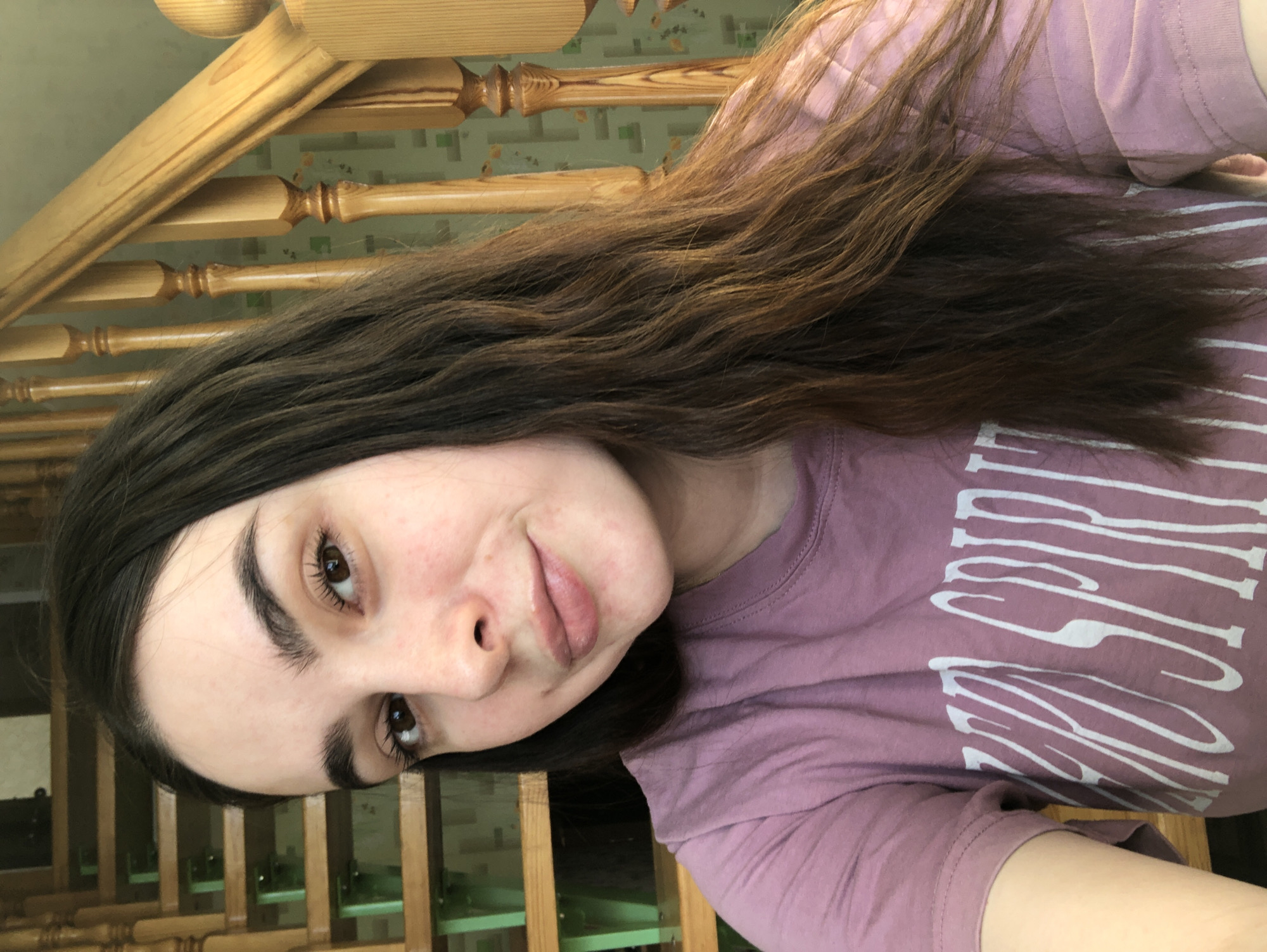 A person with brown hair and fair skin is shown in a selfie. They are wearing a mauve t-shirt with white lettering. The background includes a wooden staircase railing and patterned wallpaper. The location in the city of Noviy, Russia is not discernible from this image.