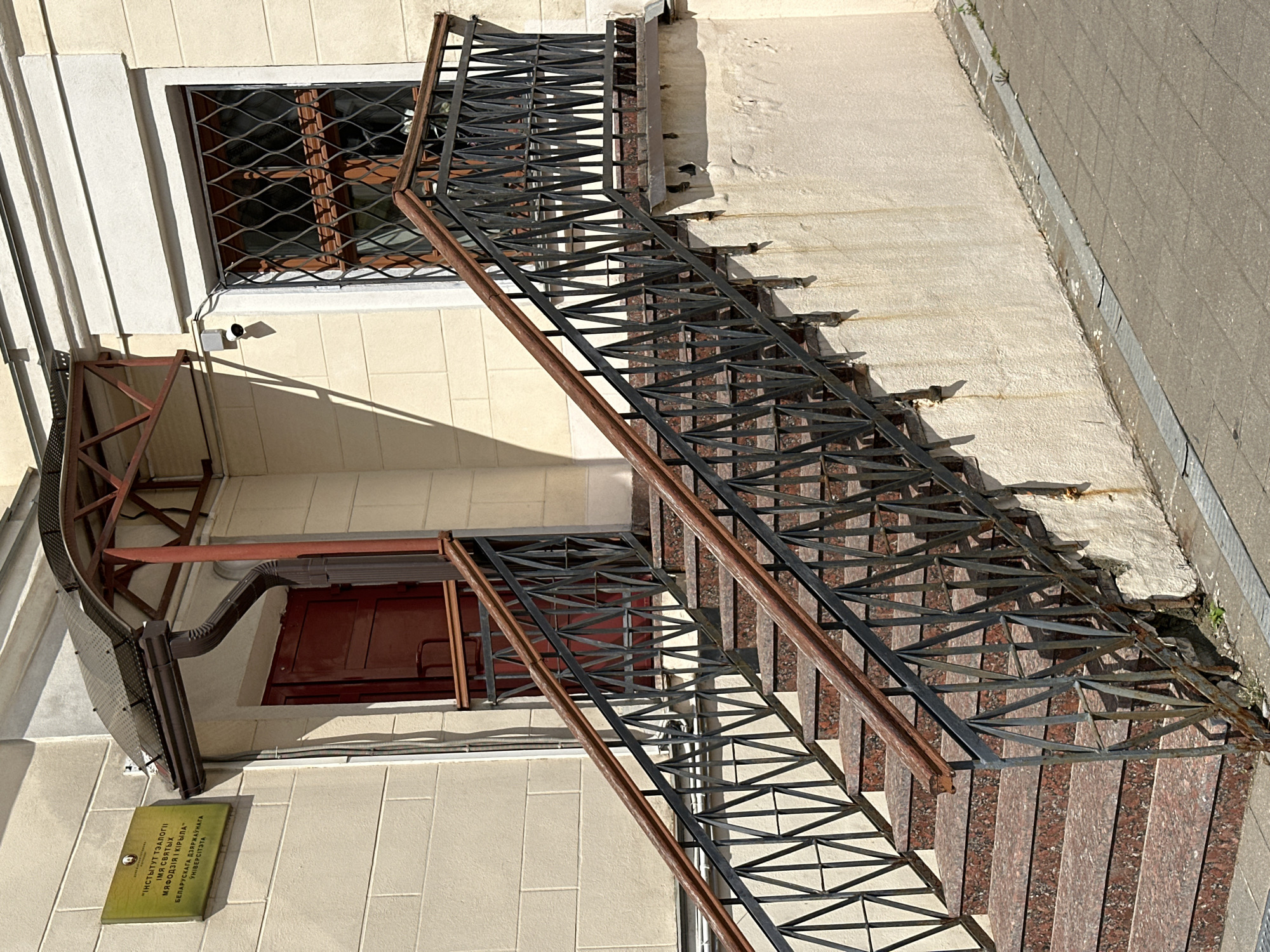 The image shows the exterior of a building with a red door and a set of stairs leading up to it. A sign on the wall above the door reads "INSTITUTE OF THEOLOGY NAMED AFTER SAINTS CYRIL AND METHODIUS OF THE BELARUSSIAN STATE UNIVERSITY". A metal railing with a decorative pattern runs alongside the stairs. A security camera is mounted on the wall near the door. The building is made of light-colored brick and has a cream-colored facade. The stairs are made of red granite and have a metal railing with wooden handrails. The scene is set on a sunny day, with shadows cast by the stairs and the building.