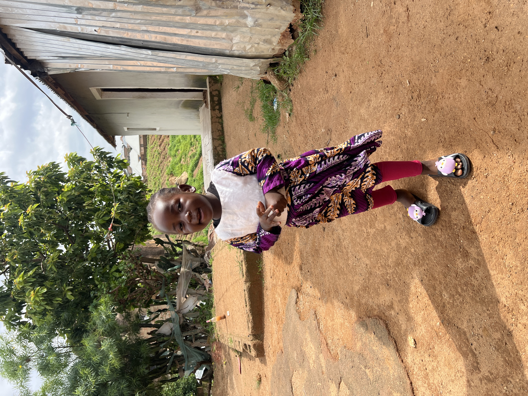 A young Black girl stands outdoors, facing the camera and smiling. She wears a white top with a colorful, patterned dress over pink leggings and black slides with cartoon character embellishments. Her hands are clasped in front of her. The background features a partially constructed building with corrugated metal siding, trees, and a dirt ground. The scene appears to be in a residential area of Jos, Nigeria.