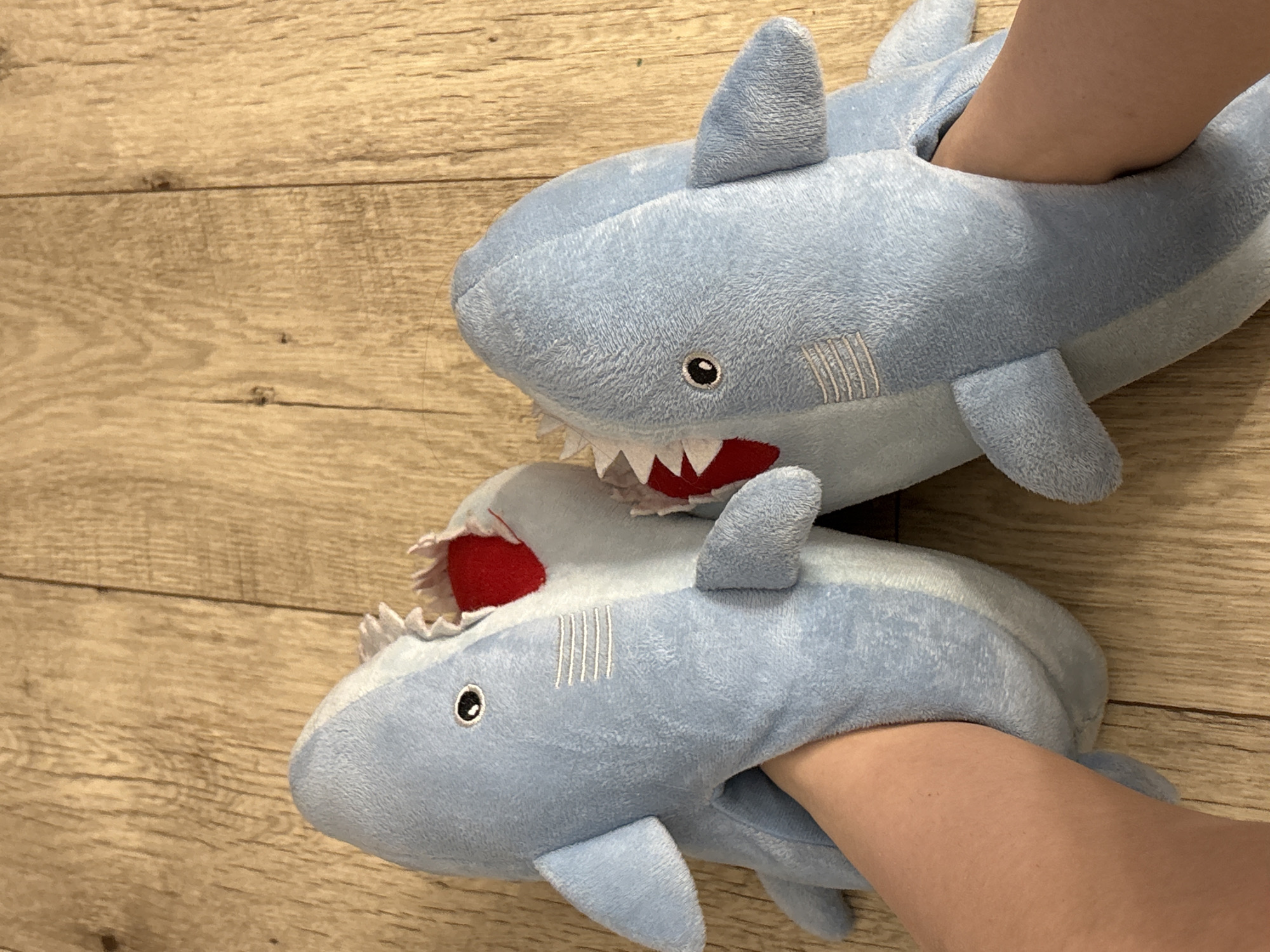 The image shows two plush blue shark slippers on a wood-grain surface. Each slipper has a white mouth with teeth and a red interior. A person's foot is inserted into each slipper. The location is not discernable from the image.