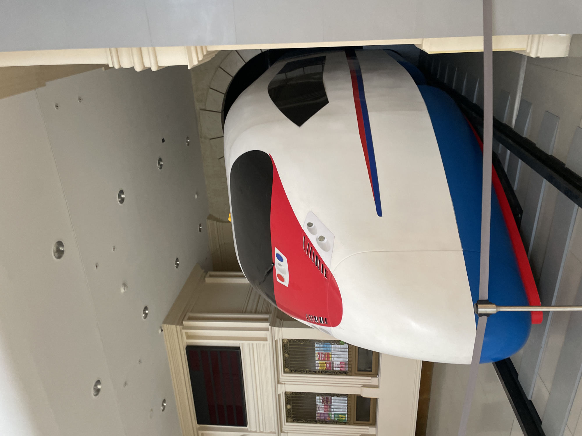 The image shows a model of a high-speed train, painted white with red and blue accents, positioned on a display track inside a building. The train is large and appears to be a static exhibit. In the background, there is architectural detailing that suggests an indoor exhibition space or museum, with columns and recessed lighting in the ceiling. To the left of the train, there are what appear to be display cases with items inside. The overall setting is clean and well-lit. The image does not contain any people, actions, or interactions. The location is not specified in the image itself.