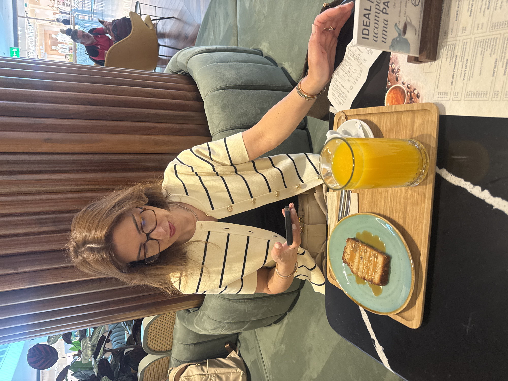 In a casual café setting in Senhora da Hora, Portugal, a woman is seated at a table, enjoying a snack and drink. She has shoulder-length brown hair and wears glasses, a cream-colored cardigan with black stripes, and a black top. Her left arm is extended towards a menu stand, while her right hand holds a dark smartphone.
On the dark-colored table with white accents, a wooden tray holds a slice of cake or bread with caramel drizzle on a light blue plate, and a tall glass of orange juice. A paper receipt is visible near the menu stand. The woman is seated on a green upholstered bench, with a light-colored bag resting on the floor beside her. In the background, wooden slatted partitions provide a dividing element, and reflections reveal other patrons, including a person in a red shirt, and furniture like a yellow armchair and rattan-style seating. Green plants add to the decor.
Visible text from the menu includes "PASTELARIA," "BOLOS A FATIA" (cakes by the slice), "BOMBONS À UNIDADE" (chocolates by the unit), and phrases like "IDEAL acompanha uma PA" (IDEAL to accompany a pastry). Prices such as "1.00€" and other numbers are also legible. The word "FALCONERE" can be seen in the background reflection. The atmosphere appears relaxed, suggesting a coffee break or casual meal.