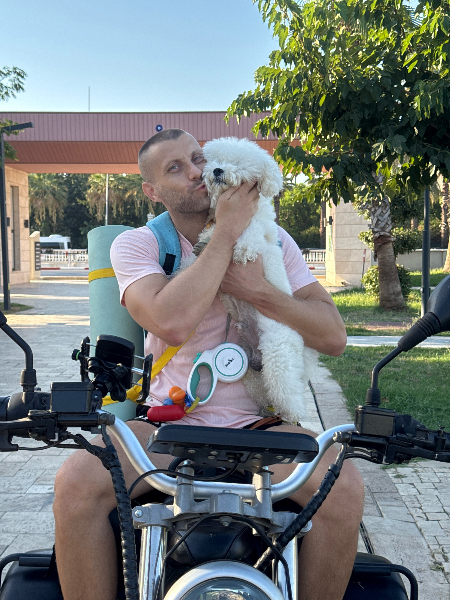 In Antalya, Turkey, a man is sitting on a motorcycle, holding a white Bichon Frise dog to his face and kissing it. He is wearing a pink t-shirt, denim shorts, and a blue backpack. A rolled-up yoga mat is strapped to his back. The dog is white and fluffy. The motorcycle is parked on a paved path, with grass and trees visible in the background. A building with a pink facade and a palm tree are also present. The sky is clear and blue.