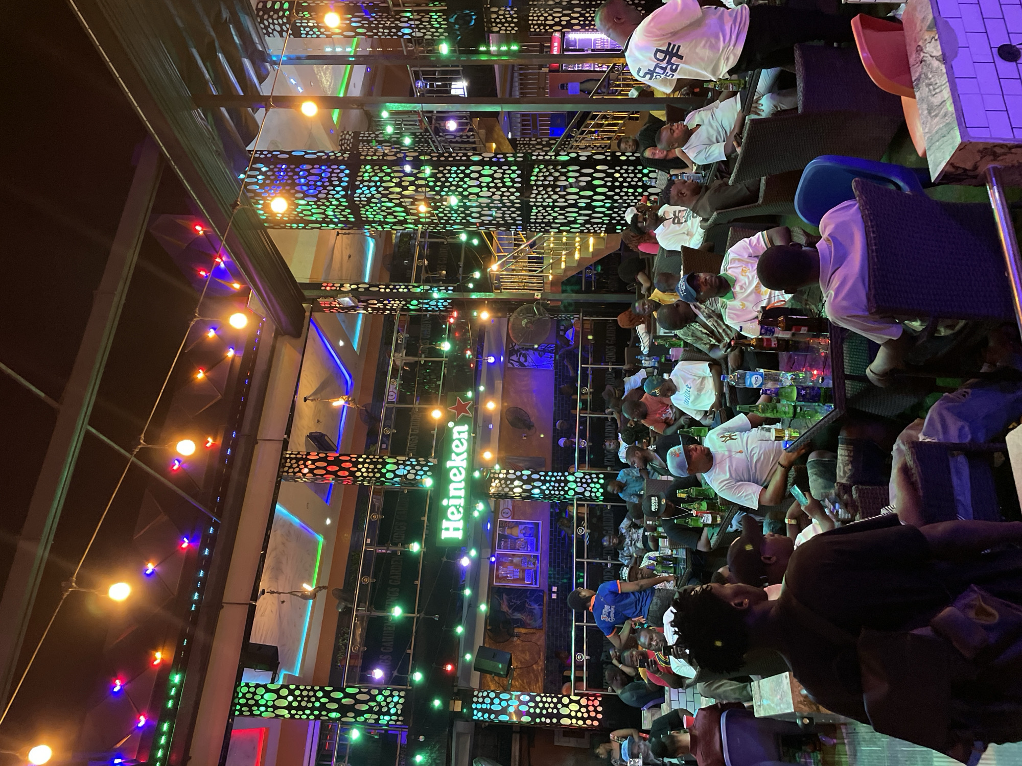 This image shows an outdoor bar in Obosi, Nigeria. Many people are sitting at tables, drinking from glass bottles. There is neon lighting and a large "Heineken" sign.