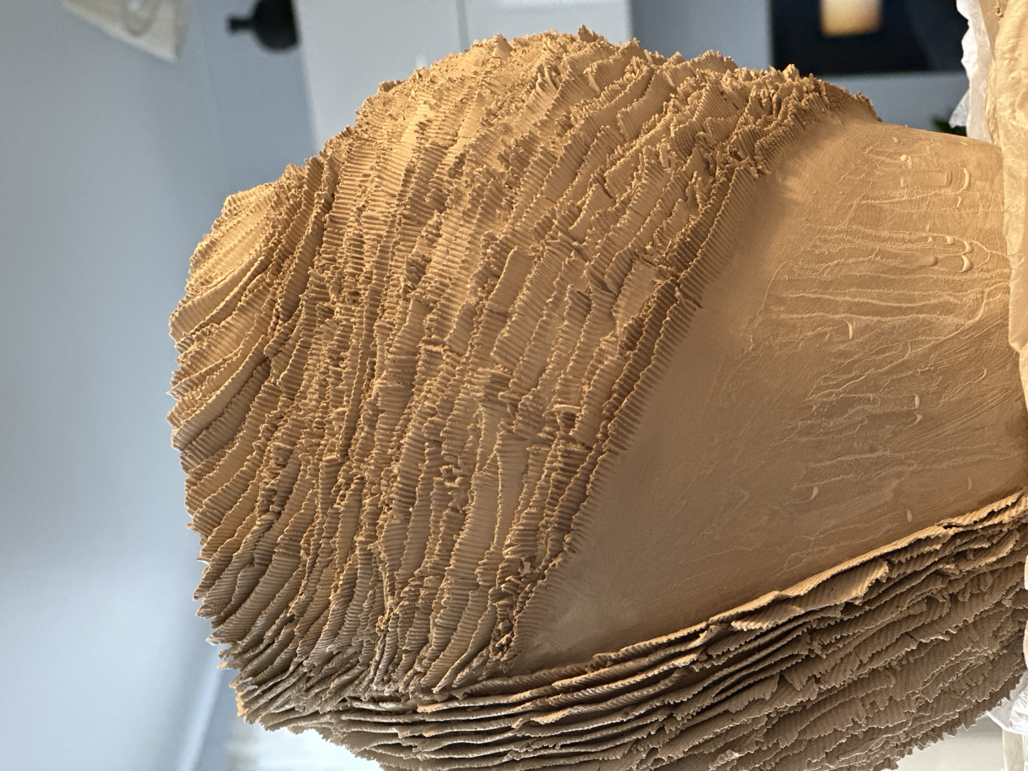 The image shows a close-up, detailed view of a large, textured, spherical object, likely a sculpture or decorative item, made of a light brown material that appears to be clay or a similar ceramic. The surface is covered in many small, parallel, vertical ridges, giving it a layered or fluted appearance. The lower portion of the object has a smoother texture with some vertical streaking. The object is resting on a white surface, possibly fabric or paper. The background is blurred, showing hints of an indoor environment with a pale blue ceiling, white curtains, and a dark object in the distance. There is no visible indication of the city of Nevinnomyssk, Russia, or any people.