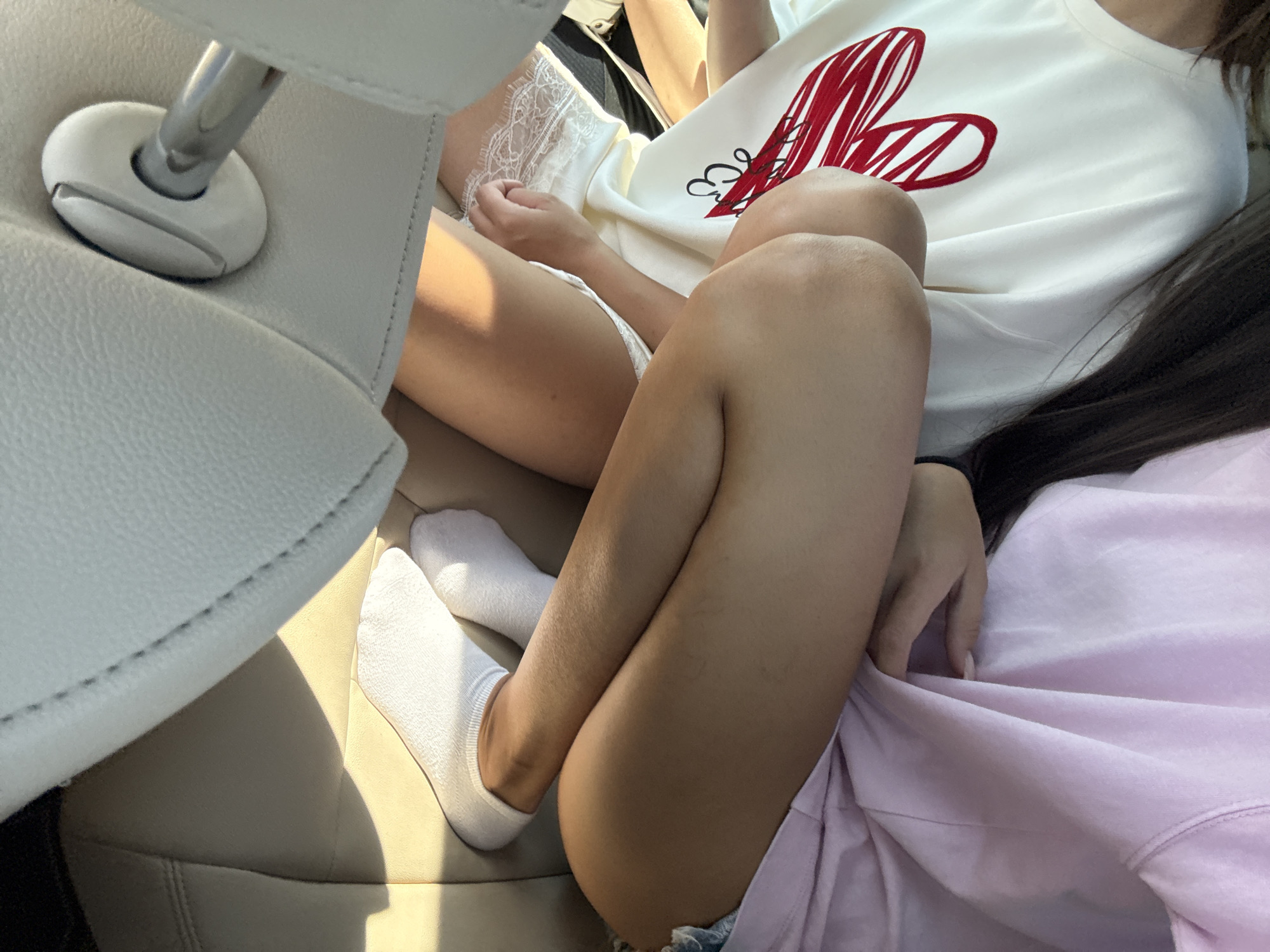 The image depicts two individuals sitting in a car, likely in Arpașu de Jos, Romania, due to the presence of a car interior commonly found in the region. The foreground shows the legs of one individual, wearing white socks and denim shorts, crossed over the legs of another individual. This second individual is wearing a white t-shirt with a large red heart design and lace trim. The car's interior features light-colored leather seats and a metal bracket attached to the seat. The lighting suggests it is daytime, with sunlight casting shadows.