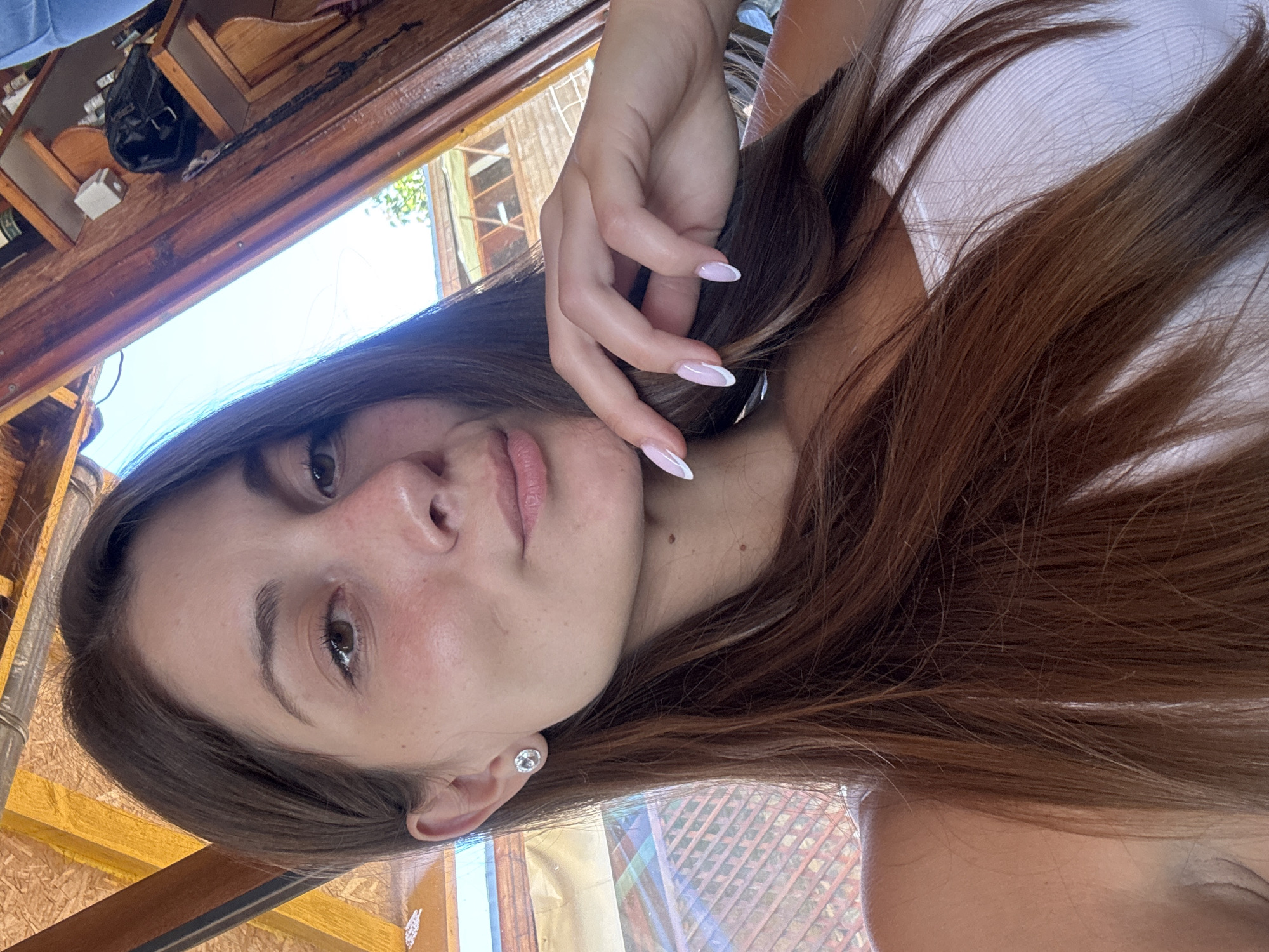 A woman with long, brown hair, wearing a white tank top, is shown from the chest up. She has a stud earring in her left ear and her nails are painted with a French manicure. Her right hand is resting on her chin. The background consists of wooden structures and a glimpse of a blue sky with greenery. The scene appears to be outdoors, possibly in Sozopol, Bulgaria, judging by the style of the wooden architecture which is typical of coastal towns in the region.