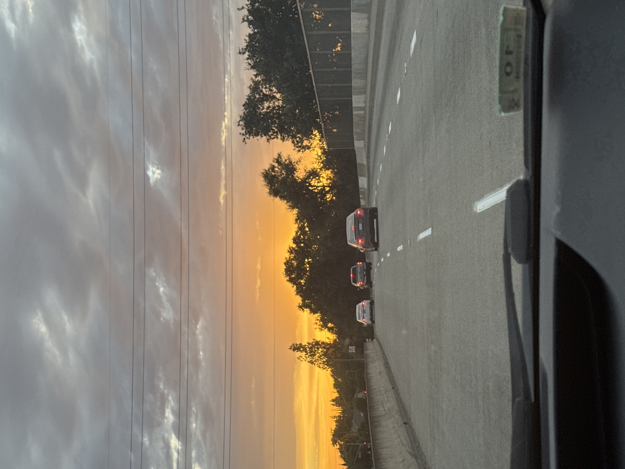 The image captures a view from inside a vehicle driving on a highway at sunset. The sky is filled with dramatic clouds tinged with orange and yellow from the setting sun. Multiple lanes of asphalt are visible, with white dashed lines marking the lanes. Several cars are driving on the highway, including a white sedan in the left lane, a silver sedan in the middle lane, and a dark sedan in the right lane. Trees and vegetation line the side of the highway, with a metal fence visible on the right. The foreground shows the interior of the car, including the dashboard and windshield wipers. Based on the available information, this scene is located on a highway in or near Foothill Farms, United States.