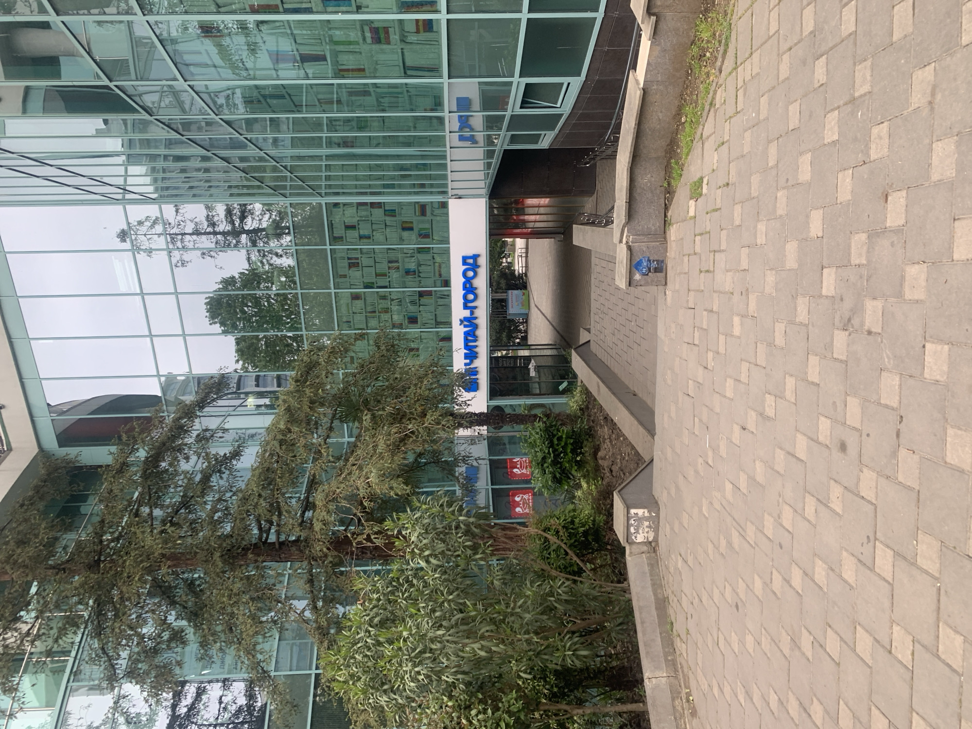 The image shows the exterior of a building with a modern glass facade, possibly a commercial or institutional building. A sign in blue lettering, which appears to be Cyrillic (ЧИТАЙ-ГОРОД), is prominently displayed above the entrance. The glass facade reflects trees and the sky, creating a semi-transparent effect. A brick walkway runs alongside the building, and a raised flowerbed with trees and greenery separates the walkway from the building's base. Two bags or merchandise with a logo are visible behind the trees. A light post is also present. The exact location within the city of Unknown, Unknown is not discernible from the image, but the style of the building and paving suggests a modern urban environment. There are no people visible in the immediate scene. The environment is characterized by the interplay of natural elements (trees, sky reflection) and urban construction.