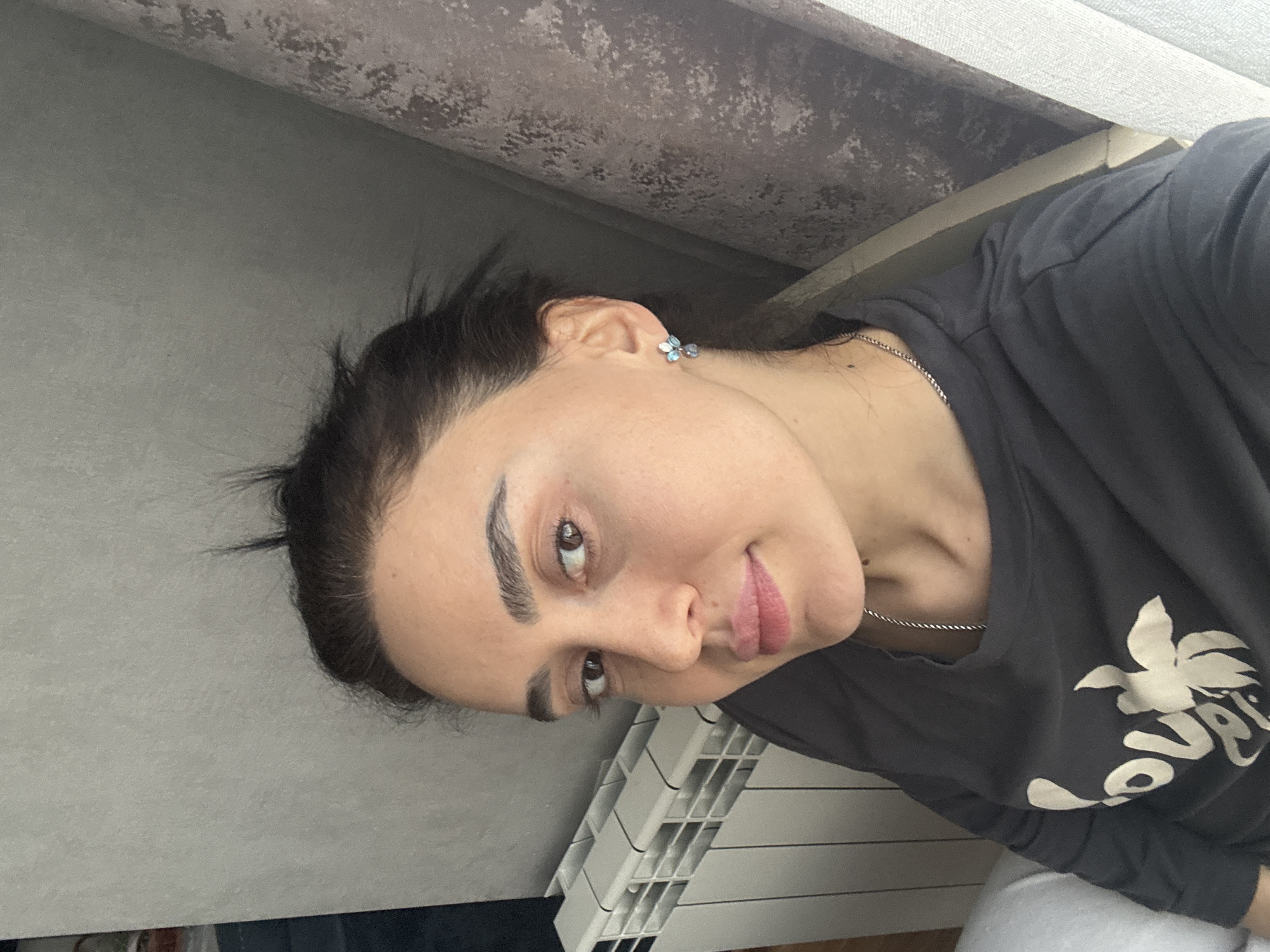 A person is captured in a close-up, off-center portrait. The individual has dark hair and fair skin, and wears a gray t-shirt with a white floral design. The person is wearing a delicate silver necklace and small flower-shaped earrings. The background shows a gray wall, a textured ceiling, and a white radiator. It is not possible to determine the location within the city of Tbilisi, Georgia from the provided visual information.