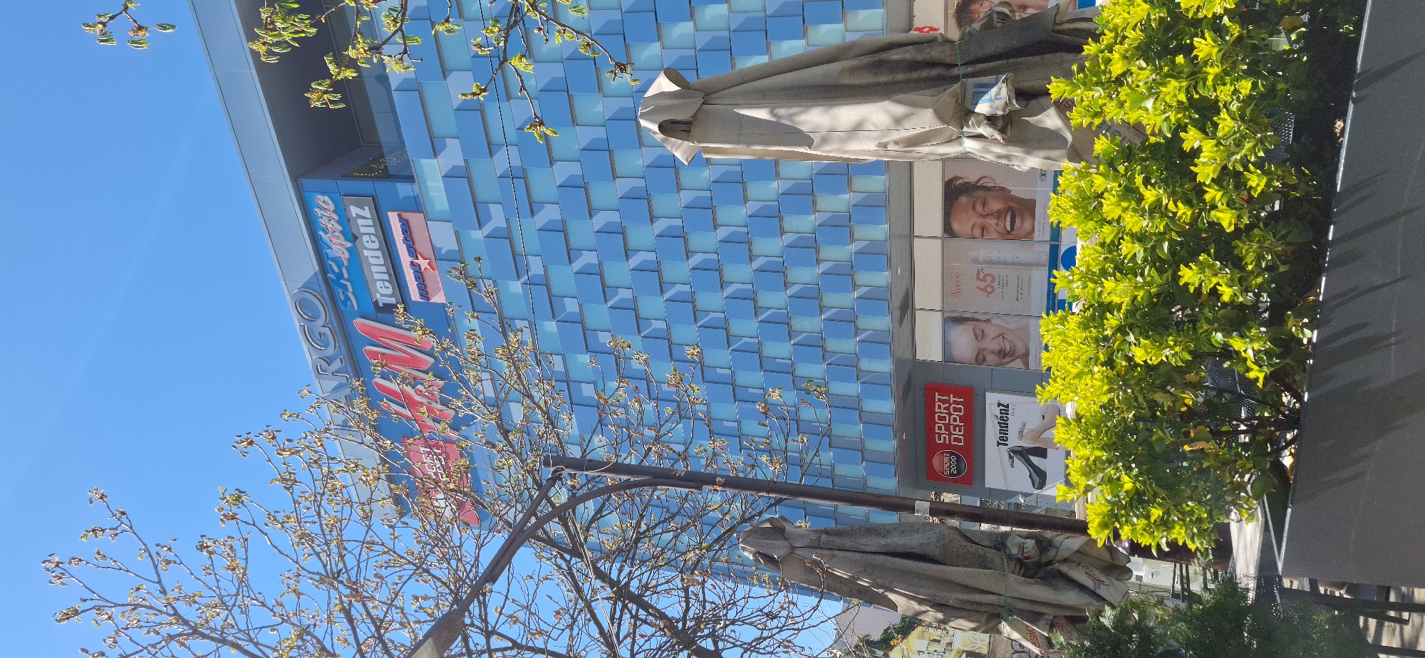 The image shows a building facade with "ARGO" and "Tendenz" signage in Blagoevgrad, Bulgaria. The building's exterior is modern, featuring a blue tiled pattern. Trees with budding leaves are in the foreground, partially obscuring the view. Umbrellas and green foliage are also visible. Advertisements, including beauty product ads, are displayed on the building's lower levels, and a "Sport Depot" sign is noticeable. The sky is clear and blue.