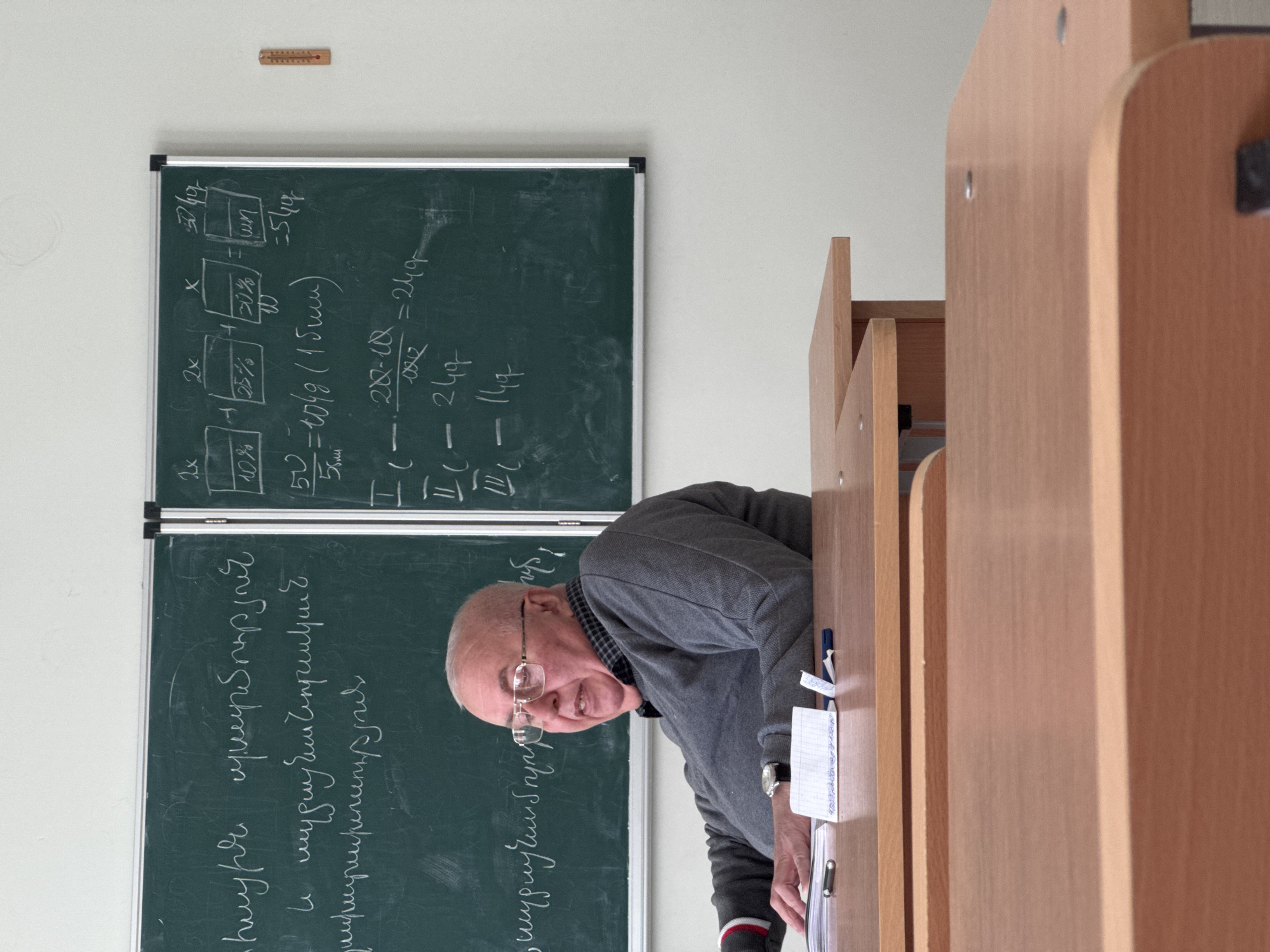 The image depicts an older man with light skin, who is bald with short white hair on the sides and wearing glasses. He is looking directly at the camera with a neutral to slightly smiling expression. He wears a grey long-sleeved sweater over a plaid shirt. The man is positioned at a light-colored wooden desk, with his left hand resting on a stack of papers and an open, spiral-bound notebook that holds a blue pen.
The setting appears to be a classroom or lecture hall. Behind and to the left of the man is a green chalkboard covered with extensive chalk writing. The writing includes both mathematical equations and text in Armenian script. The Armenian text features phrases such as "հայերեն" (Armenian), "մեր հայրենիքի պատմություն" (history of our homeland), and "Հայոց ցեղասպանություն" (Armenian Genocide). The mathematical content includes variables (e.g., "2x," "x"), percentages (e.g., "10%," "25%," "20%," "50%"), and equations like "IL - 280-10 = 249" and "50 = 104g (15mm)."
Above the chalkboard, a small, wooden-backed thermometer is affixed to the light-colored wall. Parts of other light wooden classroom furniture are visible in the foreground to the man's right. The scene is well-lit, typical of an indoor environment during the day. The man appears to be an instructor or lecturer, potentially teaching or engaging with a class. The Armenian text on the blackboard confirms the context of Yerevan, Armenia.