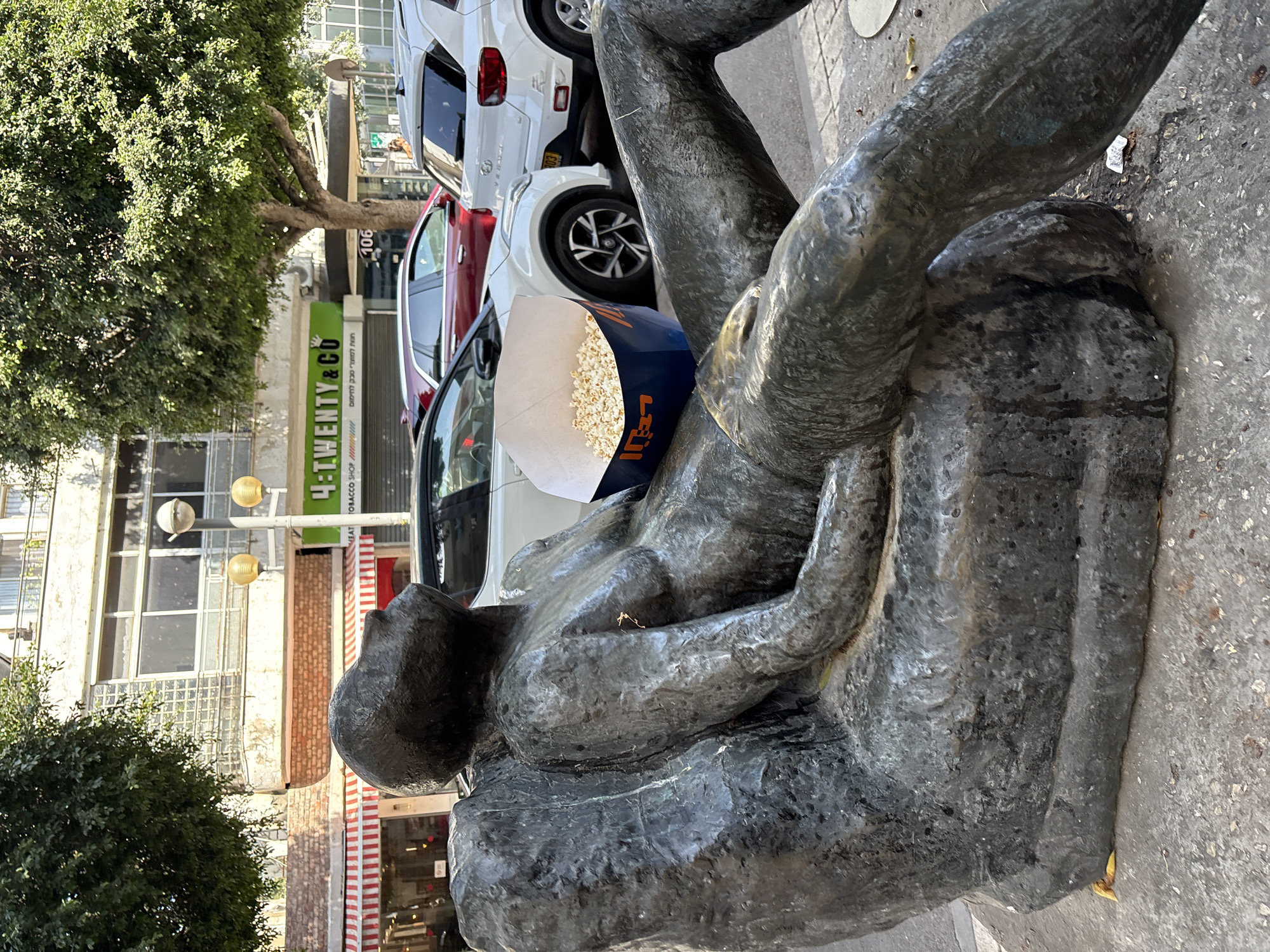 A street scene in Tel-Aviv, Israel. The focus is on a bronze statue of a reclining male figure. A box of popcorn rests on the statue's chest, with the brand name in Hebrew. Behind the statue, there are parked cars and a store sign that reads "4:Twenty & Co TOBACCO SHOP." The overall atmosphere suggests a public space with commercial establishments nearby.