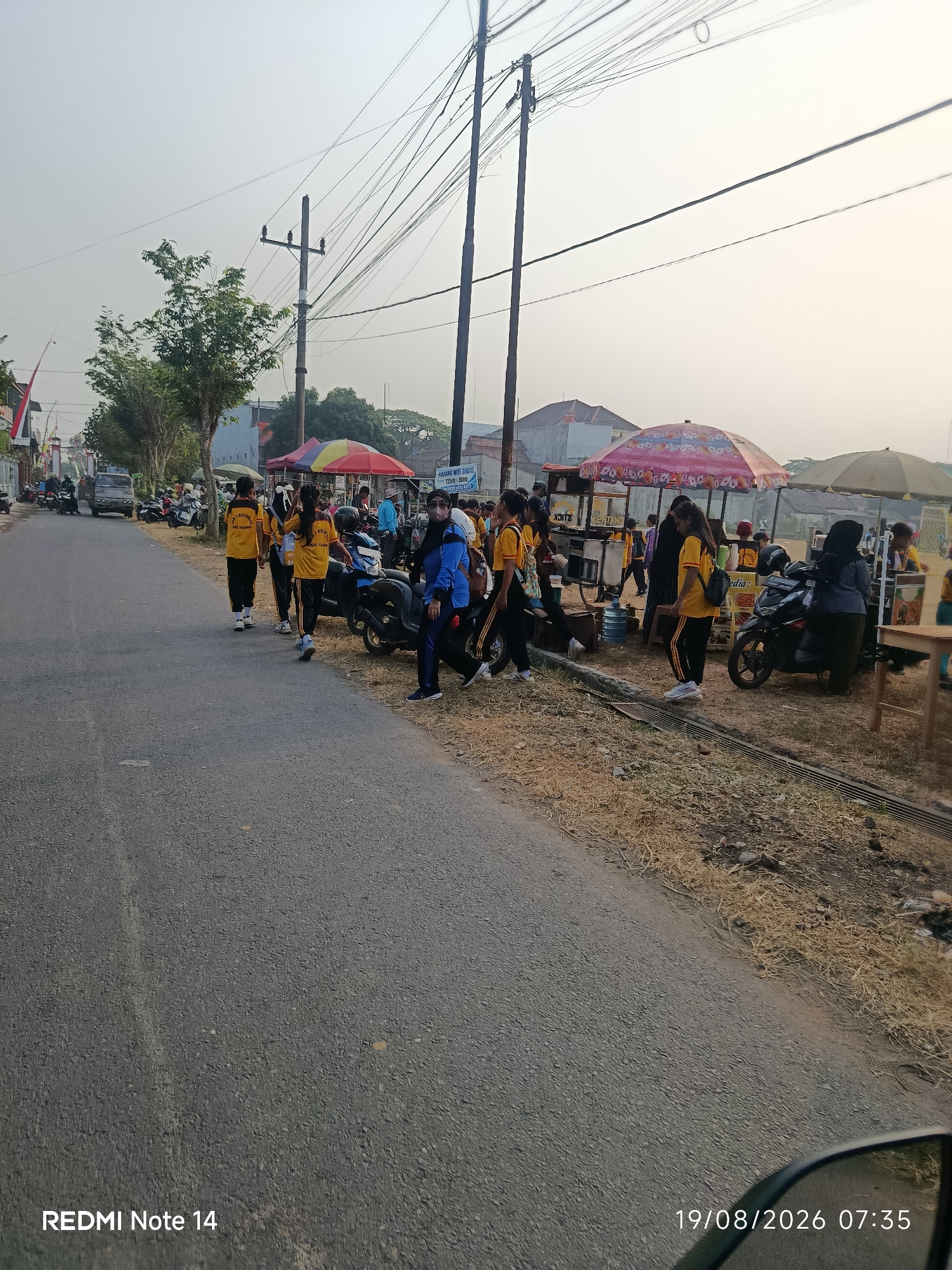The image depicts a street scene in Panggungrejo, Indonesia, on a clear day with bright sunlight. A paved road occupies the foreground, with a dry, grassy embankment on the right side. A group of individuals, predominantly young people wearing yellow shirts and black pants, are walking along the road and gathered near parked motorcycles and food stalls. Several food stalls with umbrellas are visible, suggesting a market or event. Motorcycles are parked along the roadside, and a water dispenser is present near one of the stalls. The background shows buildings, trees, and utility poles with numerous overhead wires. The overall atmosphere appears casual and communal.