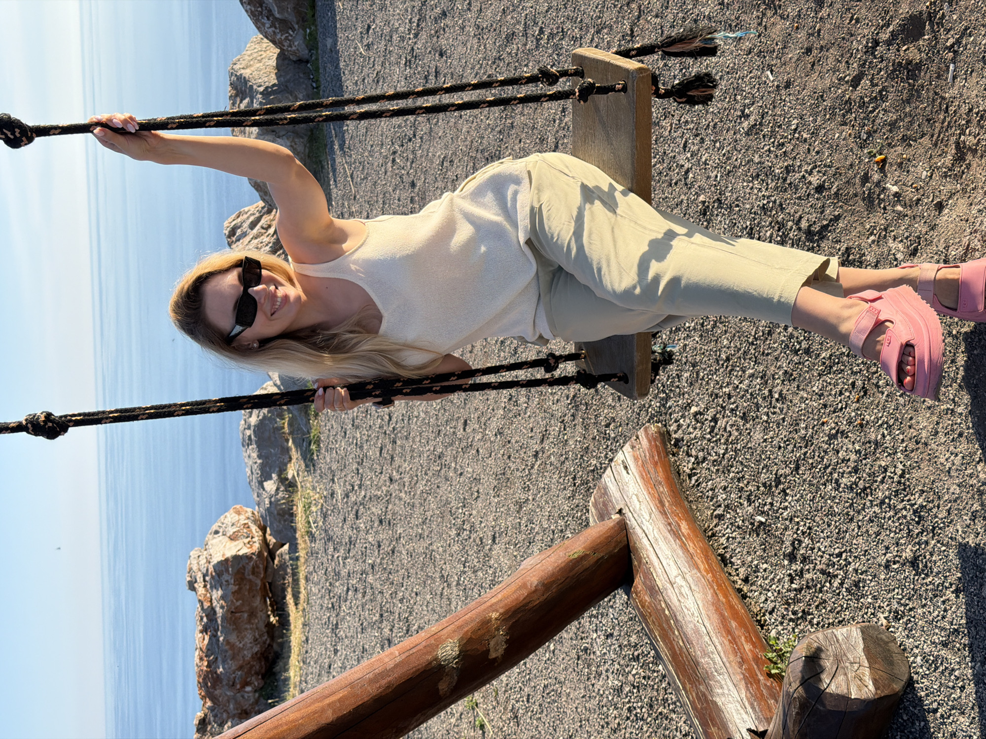 A blonde woman with sunglasses is sitting on a wooden swing outdoors. She is wearing a cream-colored tank top and light green pants, with pink platform sandals. The swing is suspended by thick ropes from a wooden frame. The background shows a body of water, possibly the Adriatic Sea, with a clear blue sky above. Rocky formations and gravel cover the ground. The scene appears to be set in a coastal area, potentially in Kovačko Polje, Montenegro, given the hint.