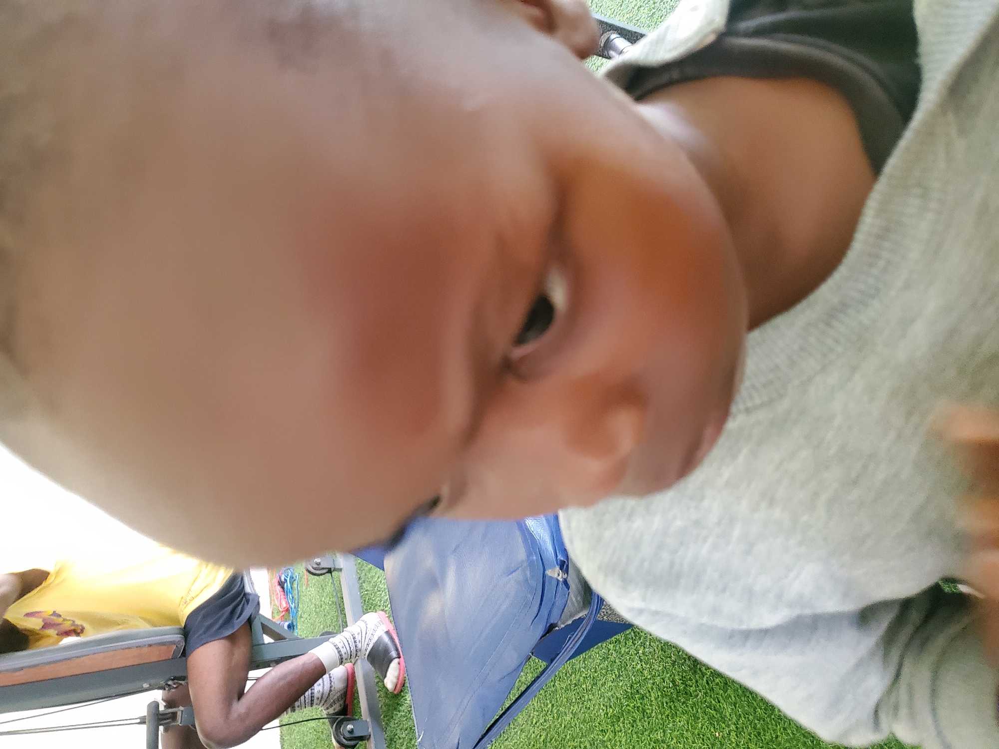 In Jos, Nigeria, a close-up shot focuses on a young child's head and upper body. The child is wearing a light grey long-sleeved shirt over a darker garment. The background reveals a person in a yellow t-shirt and black shorts, seated on a gym machine. This person is wearing white socks with a patterned design and black slides with pink accents. A blue seat is visible to the child's right, situated on a green artificial turf surface. The overall scene suggests an indoor or outdoor recreational area, possibly a gym or a park.