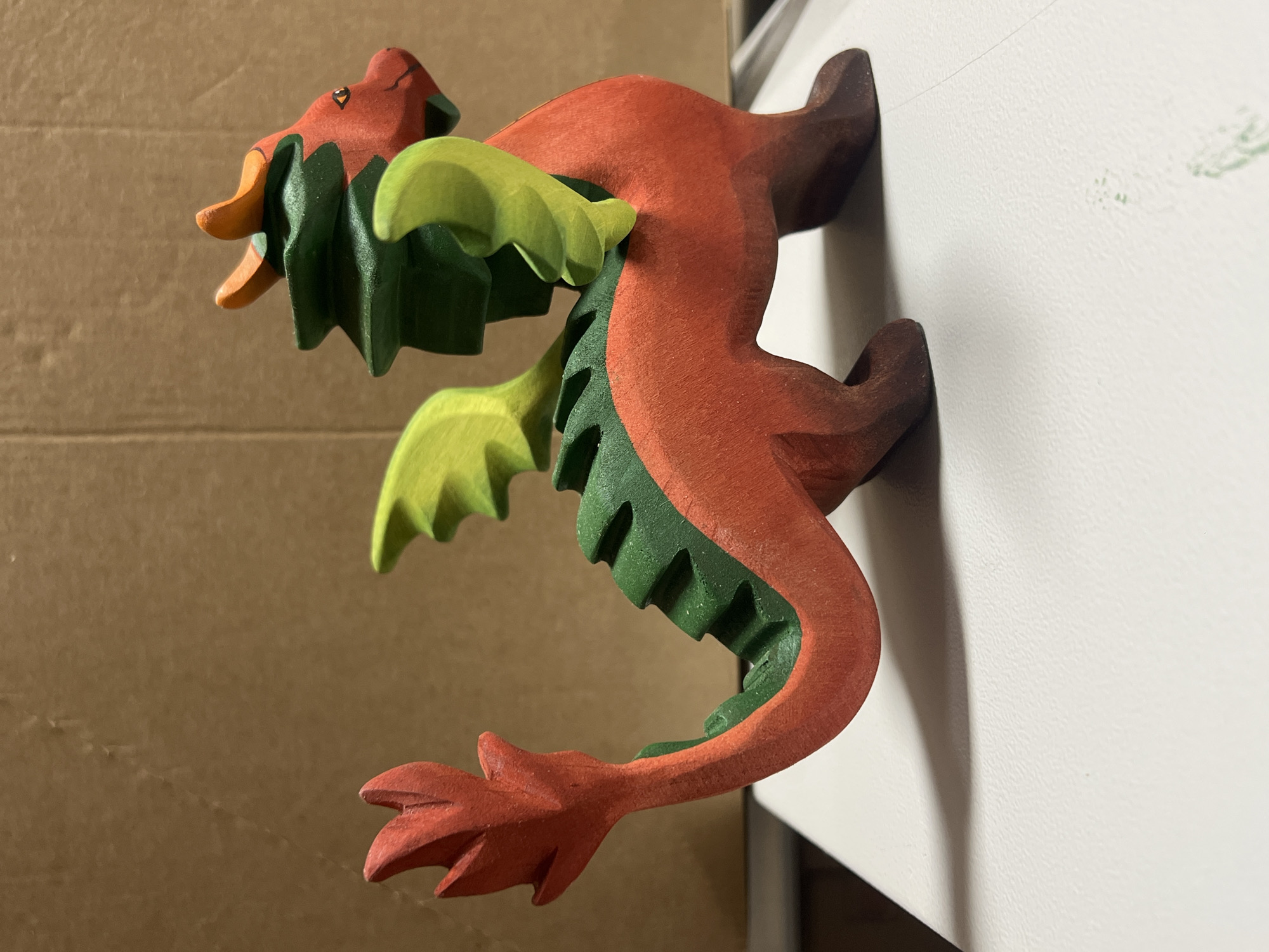 This is a close-up, side-profile shot of a wooden dragon figurine. The dragon is predominantly red, with green accents on its back and wings, and orange details around its head. It appears to be leaping or flying, with its body curved and its tail extended. The dragon is mounted on a white surface, which is casting a shadow on the right side of the frame. The background on the left is a textured brown surface, possibly cardboard or wood. The lighting suggests it is indoors, with a soft light source coming from the front and slightly from the left. The figurine appears to be a toy or a decorative item, crafted with a stylized, angular design. There are no people visible, and no text is present in the image. The focus is sharp on the dragon, while the background is slightly blurred, drawing attention to the subject.