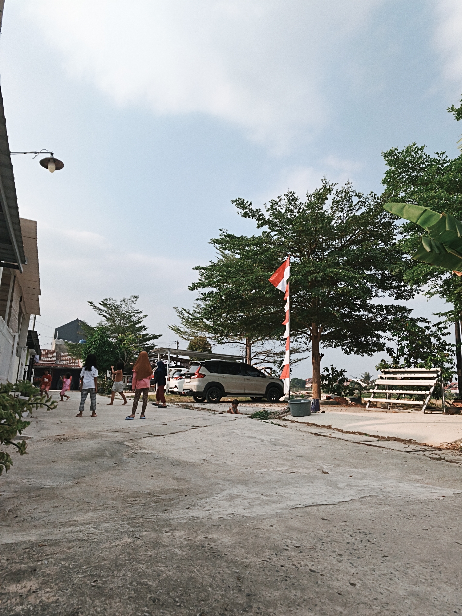 The image depicts a street scene in Bandar Lampung, Indonesia, characterized by a cloudy sky and warm daylight. The foreground is dominated by a concrete pathway with visible cracks and wear. On the left side of the pathway, a building with a tin roof and white walls is partially visible, with a metal lamp fixture attached to its exterior. A group of approximately six individuals, appearing to be children and young women, are scattered across the pathway, some engaged in activity. One person in the center is wearing a hijab and a pink top and patterned leggings. To the right of the pathway, a white multi-purpose vehicle is parked. Further to the right, a large tree with lush green foliage stands, adorned with a red and white flag on a pole. Next to the tree is a white tiered shelving unit, possibly for plants or storage. Beyond the immediate area, more trees and buildings are visible in the background, suggesting a semi-urban or residential environment. The overall atmosphere is casual, with a sense of everyday life unfolding outdoors.