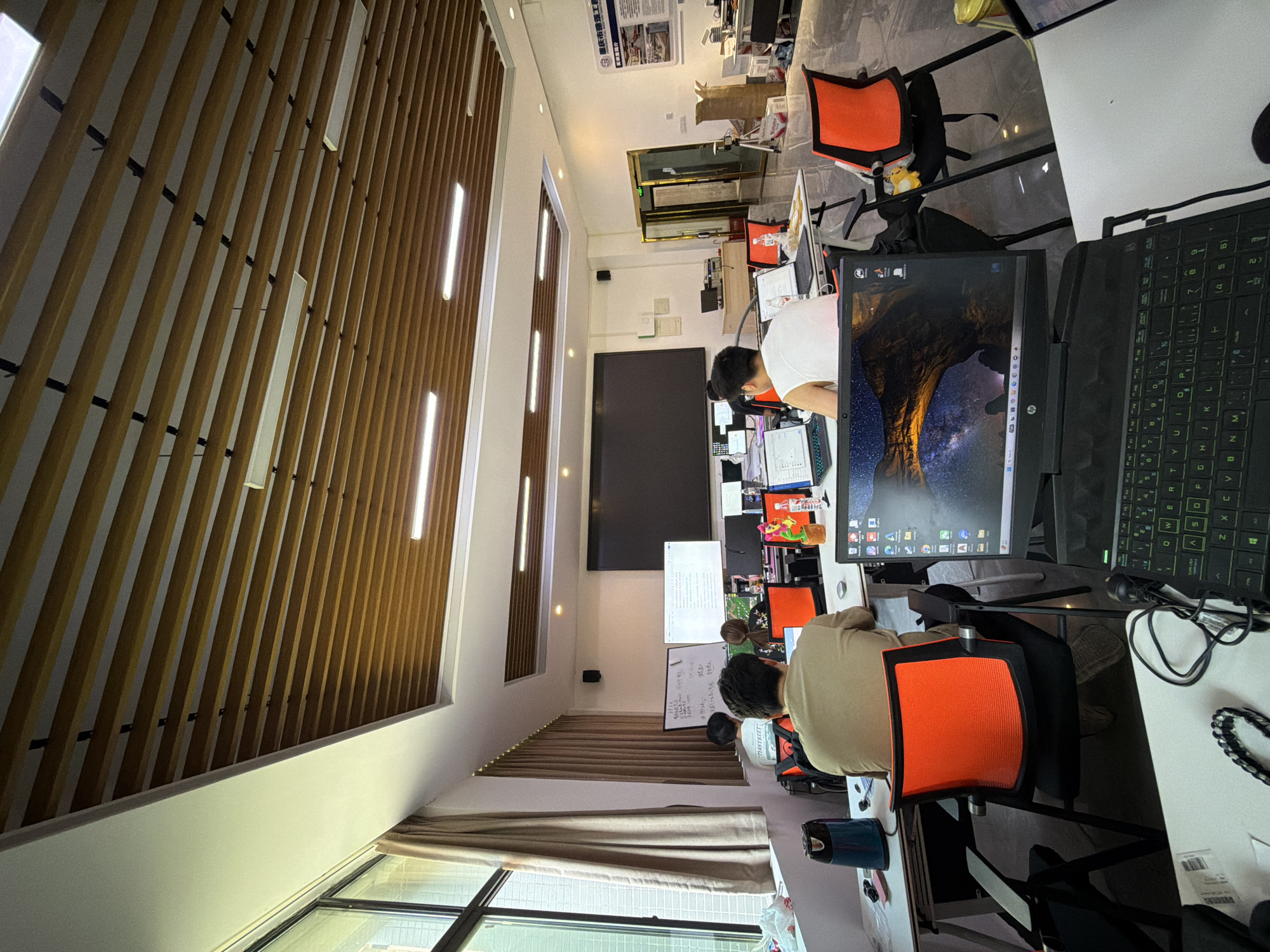 The image shows a classroom or co-working space in Jiangjin, China, with a large screen at the front, whiteboards, and multiple desks with computers. Several people are seated at desks, appearing to be working or studying. The room has a modern design with a slatted wooden ceiling and fluorescent lighting. The overall atmosphere suggests an educational or collaborative work environment.