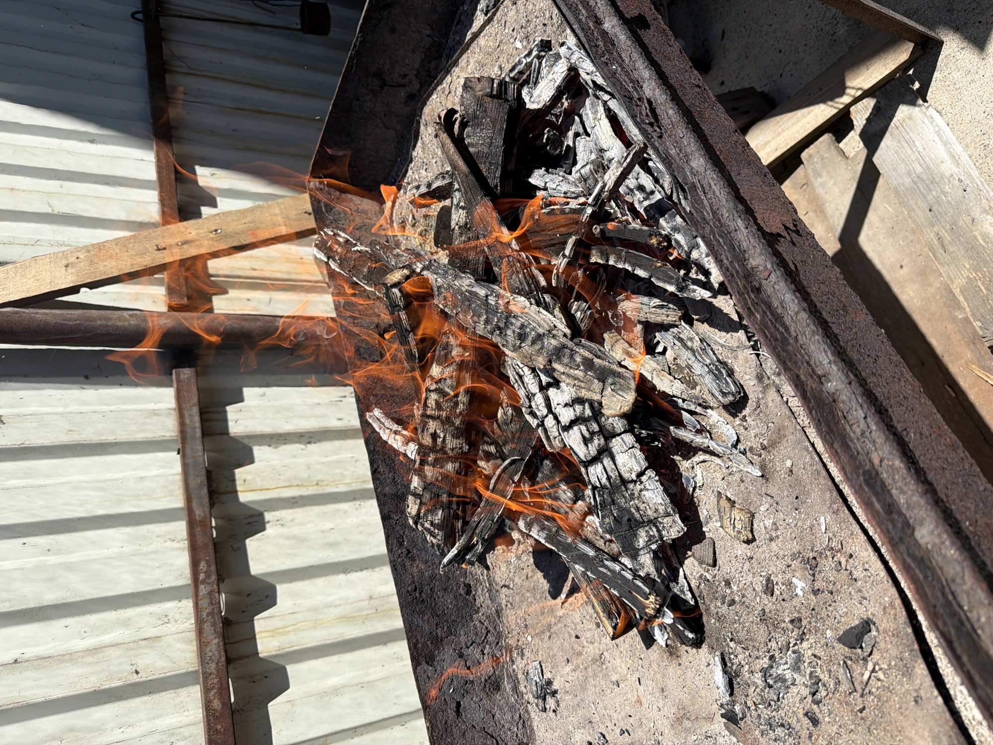 A fire burns in a metal brazier, filled with charred wood and ash. The brazier sits on a concrete surface, with wooden planks visible beneath it. In the background, a corrugated metal fence casts long shadows from the bright sunlight. The scene appears to be outdoors, possibly in a backyard or a designated outdoor cooking area. The city of Tuapse, Russia, is not explicitly depicted or suggested by any elements in the image.