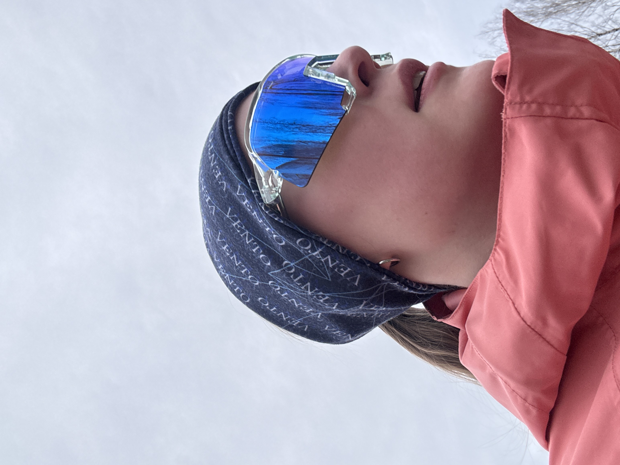 A low-angle close-up shot focuses on a person from the chest up. The individual wears blue reflective sunglasses with clear frames and a blue headband with the word "VENTO" printed on it repeatedly. A light-red or pink jacket is visible. The background is an overcast sky. The image may have been taken outdoors. There is no information available in the provided image to determine if the image was taken in the city of Chaikovsky, Russia.