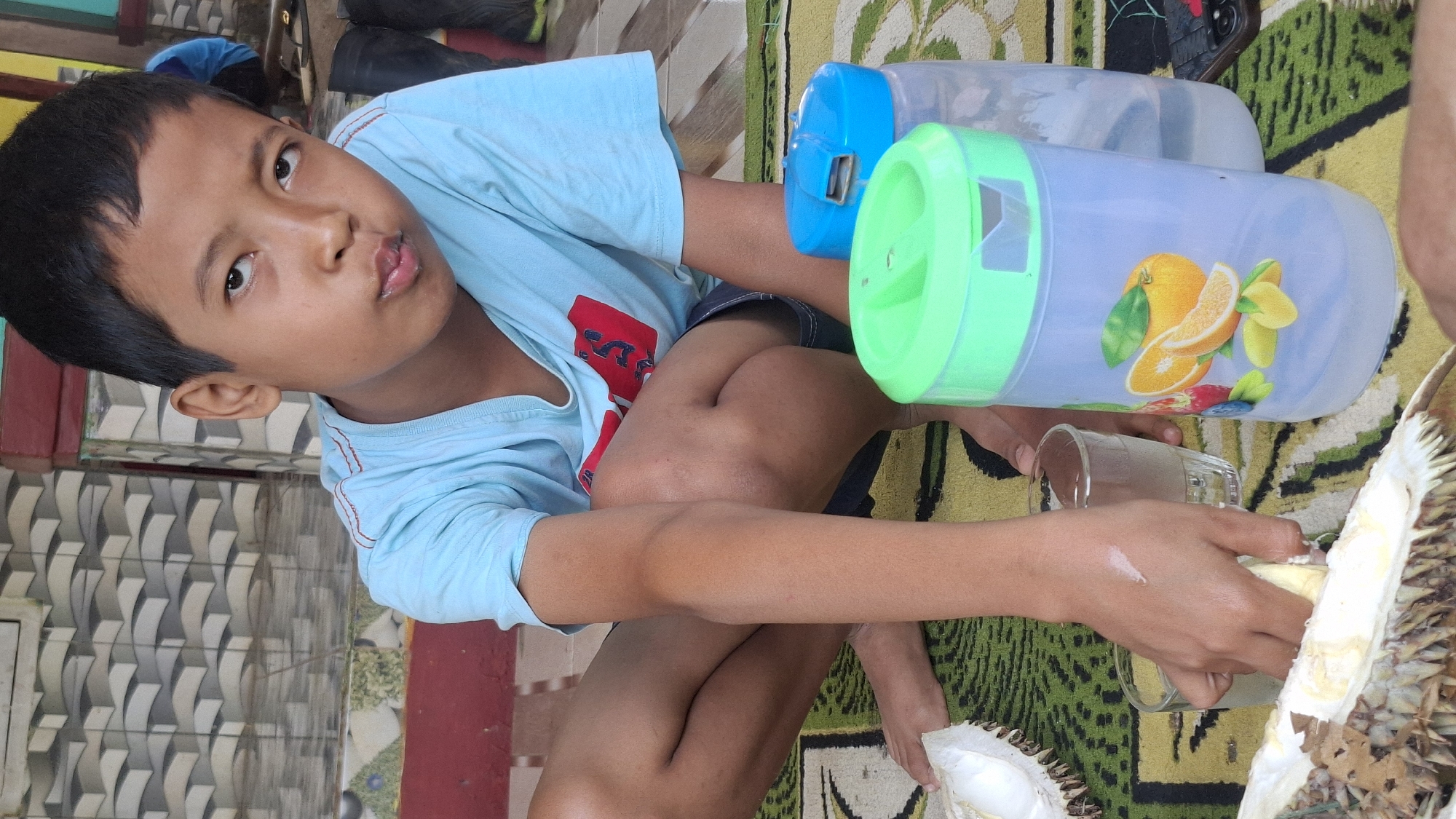 In Cilaku, Indonesia, a young boy with short black hair and a light blue t-shirt squats on a patterned rug. He is looking towards the camera with a slightly pursed expression. His right hand is reaching into a glass of clear liquid. In front of him lies a cut durian fruit, revealing its thorny exterior and creamy white flesh. To his right are two plastic pitchers: one is transparent with a green lid adorned with images of oranges and other fruits, and another is blue. A small glass of clear liquid sits between the boy's hand and the pitchers. The background features a tiled wall and a red ledge.