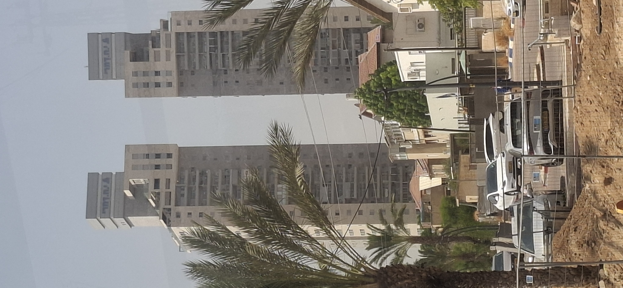 Two identical, modern high-rise apartment buildings with a stepped facade and large windows dominate the upper portion of the image. Blue Hebrew lettering is visible on the top of each building. In the foreground, several cars are parked on a street, including a white sedan and a silver SUV. Palm trees and other foliage are interspersed throughout the scene, and a construction site is visible in the lower part of the image. The buildings appear to be located in a city, possibly Be'er Sheva, Israel, based on the Hebrew script. The sky is clear and pale blue, suggesting a sunny day.