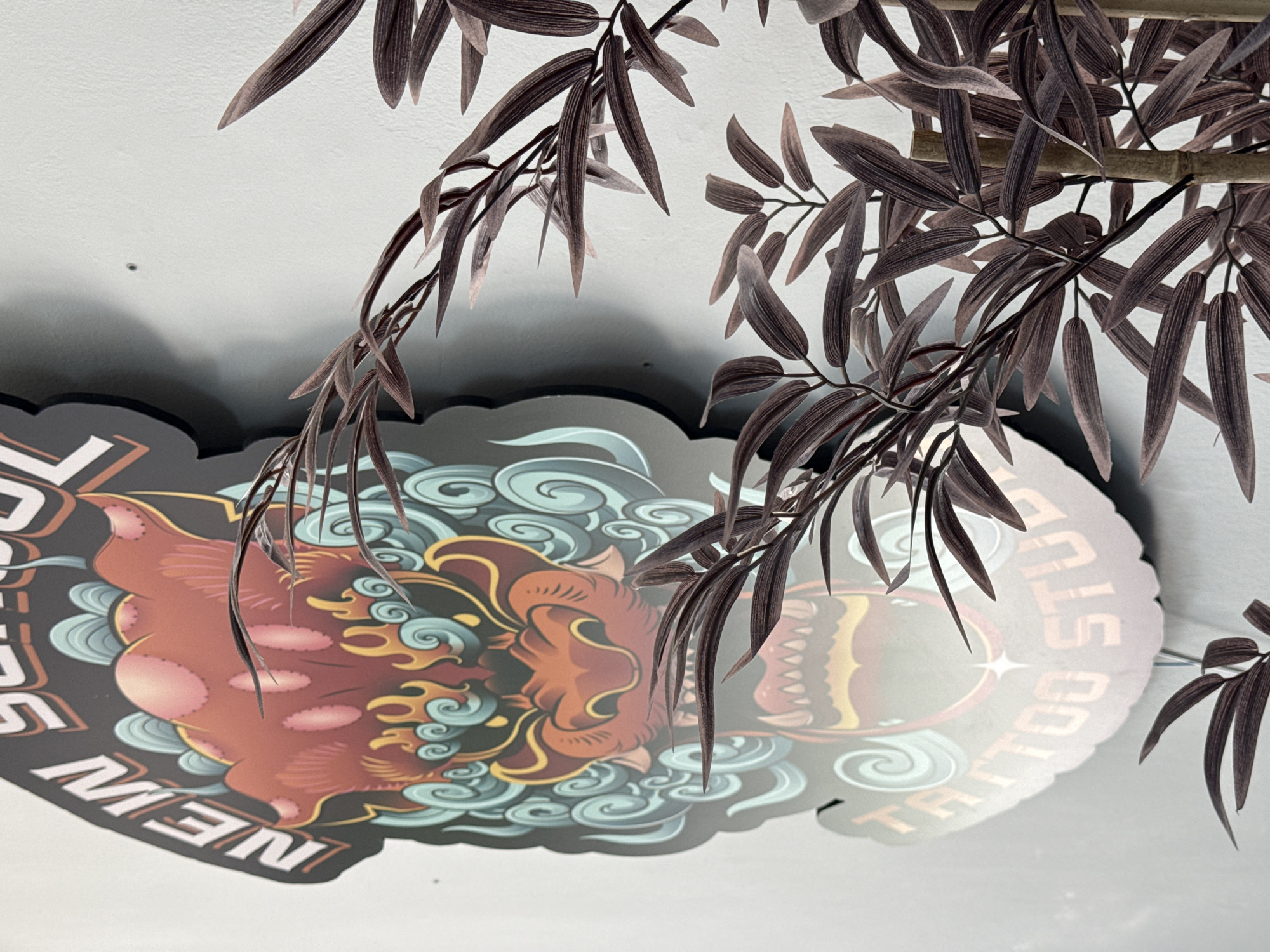 This media file captures a close-up view of a tattoo studio sign, partially obscured by artificial bamboo-like branches with dark brown leaves. The sign itself is shaped with decorative scalloped edges and features a colorful illustration. Dominating the center of the sign is a stylized representation of a mythical creature, possibly a lion or dragon, depicted in shades of red and orange with yellow accents. Swirling patterns resembling clouds or stylized waves in teal and blue surround the creature's head. Visible text on the sign includes "NEW" in white block letters on the left side, and "TATTOO STUDIO" in orange letters curving around the right side. The background is a plain, textured white surface, likely a wall or ceiling. The lighting suggests an indoor environment, possibly during the day, with a shadow cast on the white surface from the sign and branches, indicating a light source from above and to the left. The overall impression is one of a vibrant and perhaps slightly edgy establishment, with the artificial foliage adding a touch of decorative flair. The location context of Campillo Del Moro, Spain, does not provide specific visual cues within the image itself.