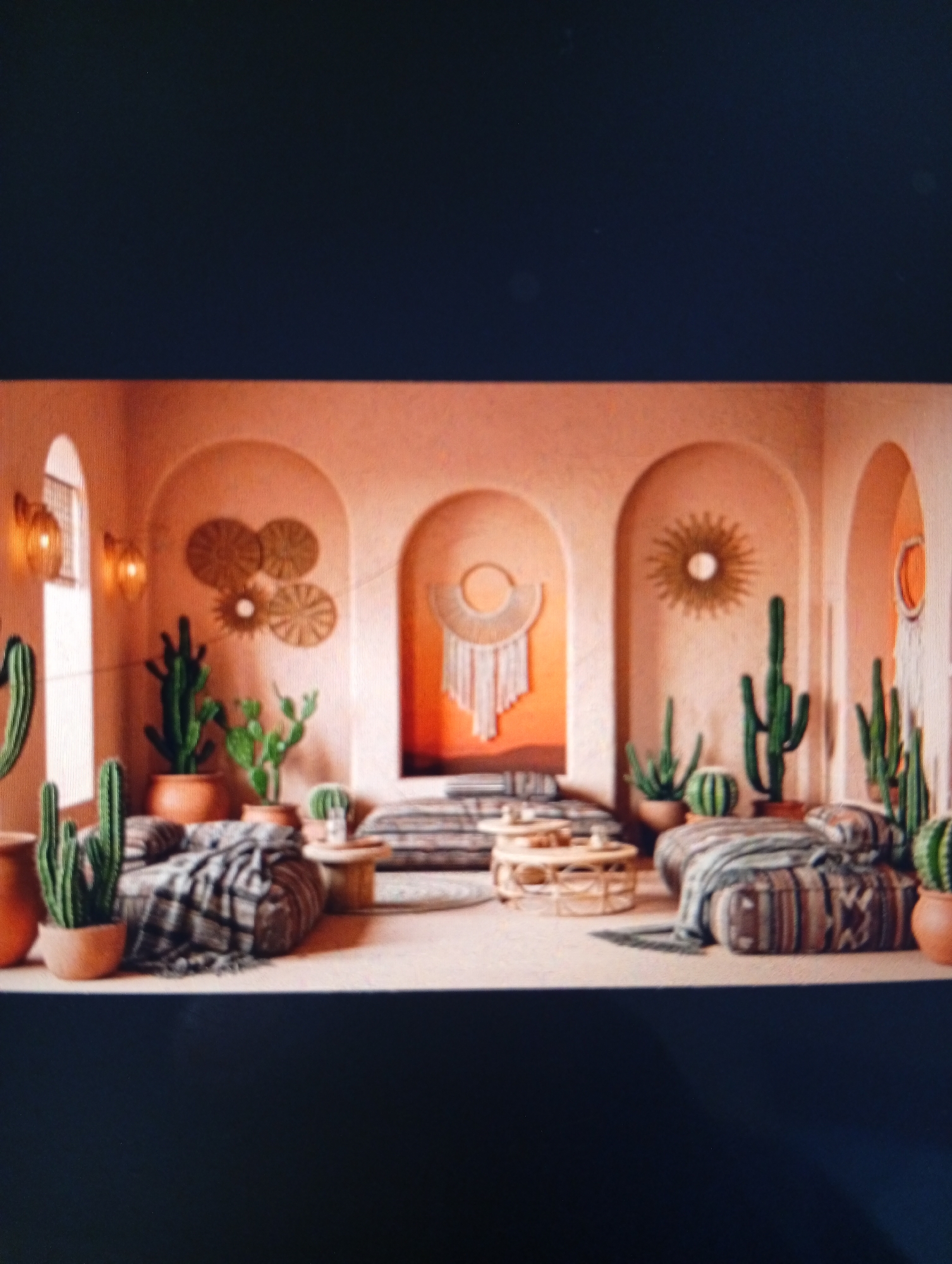 The image depicts an interior space designed with a desert or Southwestern theme. The walls are painted in a warm, earthy orange hue, with arched doorways or alcoves that create depth and visual interest. Various cacti in terracotta pots are scattered throughout the room. Seating areas include floor cushions and low benches covered in patterned textiles. Decorative elements such as woven wall hangings and sunburst mirrors adorn the walls. Small, round wooden tables serve as additional surfaces. There is no indication of a location in the city of Dutsen Alhaji, Nigeria, or any other specific location.