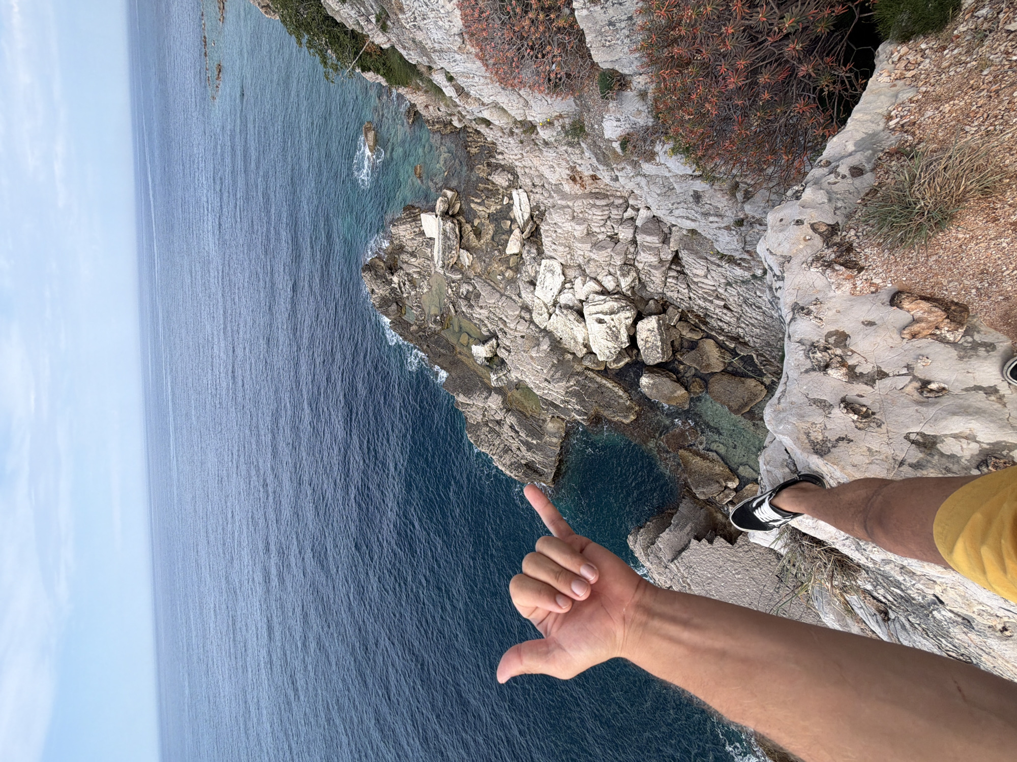 A first-person perspective photograph shows a person standing on a rocky cliff overlooking the Adriatic Sea. The location is Mirište, Montenegro. The person's left arm is extended, with their hand forming a thumbs-up gesture. The person is wearing yellow shorts and black and white sneakers. The rocky cliff is covered in sparse vegetation. The sea is a deep blue color. The sky is light blue with a few clouds.