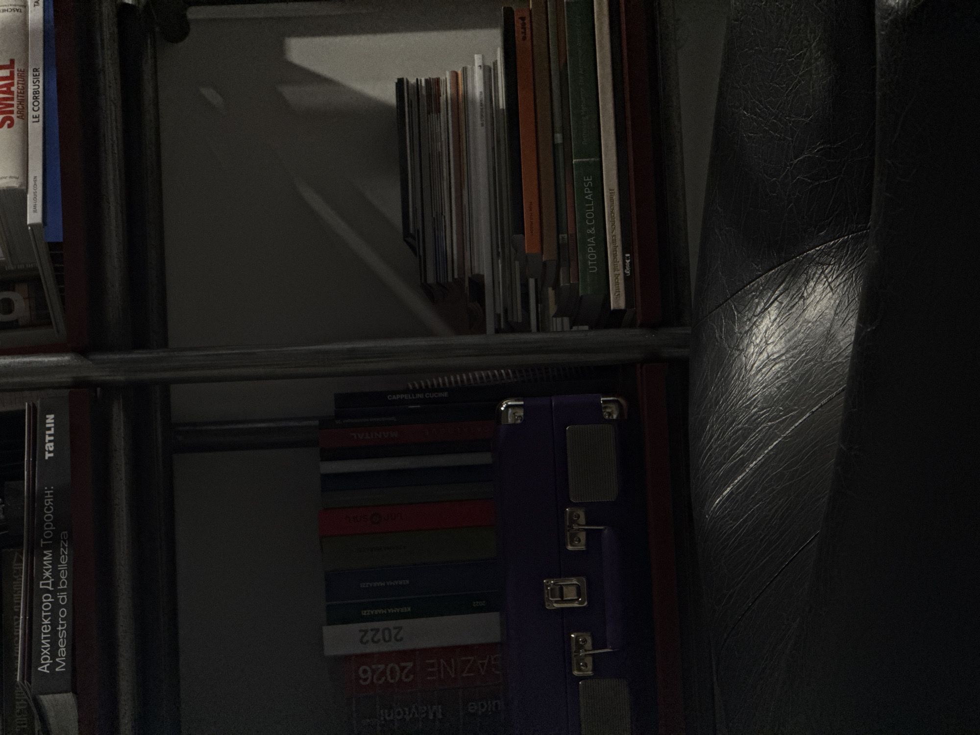 A close-up shot shows a bookshelf with several books and a dark gray leather-like object, possibly a chair. The books are stacked horizontally and vertically, with titles in multiple languages including English, Russian, and Italian. Some visible titles are "SMALL ARCHITECTURE LE CORBUSIER," "UTOPIA & COLLAPSE," "CAPPELLINI CUCINE," "MANITAL," "TATLIN," "GAZINE 2026," and "2022." A small purple suitcase-like object with metal latches is also present. The setting appears to be indoors, potentially in a study or library. There is no information in the image to suggest a specific location such as Yerevan, Armenia.