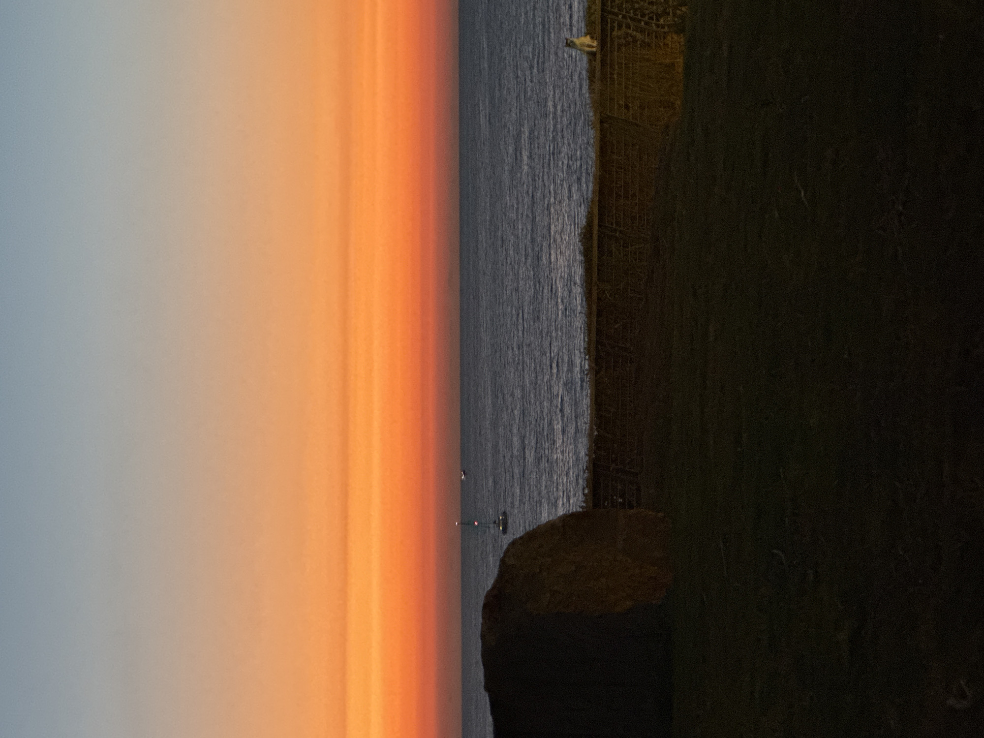 The image depicts a coastal scene at dusk, possibly in Tel Aviv, Israel, given the provided context of the question, though the location cannot be definitively confirmed from the image alone. The sky transitions from a pale blue at the top to a vibrant orange and red gradient near the horizon, indicating sunset or sunrise. The sea is a deep blue with gentle waves. In the foreground, there is a large, round hay bale on the left side, silhouetted against the sunset. A low stone wall runs horizontally across the mid-ground. On top of this wall, to the right, a slender cat is perched, facing the sea. In the distance, on the water, a few small boats are visible. The overall lighting suggests the end of the day.