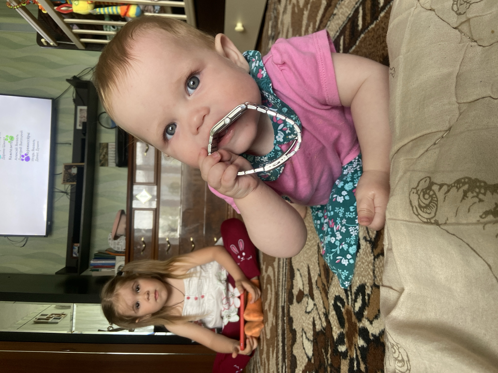 The image captures two young girls in an indoor setting. The main focus is on a baby with blue eyes and light hair, wearing a pink top with a green floral pattern trim. The baby is holding a silver watch in her mouth, looking directly at the camera. In the background, a slightly older girl is visible wearing a white top and red pants adorned with bunny images. She is holding a red phone. The surroundings reveal a patterned brown and beige carpet and a beige-colored object, possibly a couch or blanket. A television screen showing text in a foreign language is in the background. Furniture is also visible, including a crib. Based on the metadata, the location is Фоки, Russia.