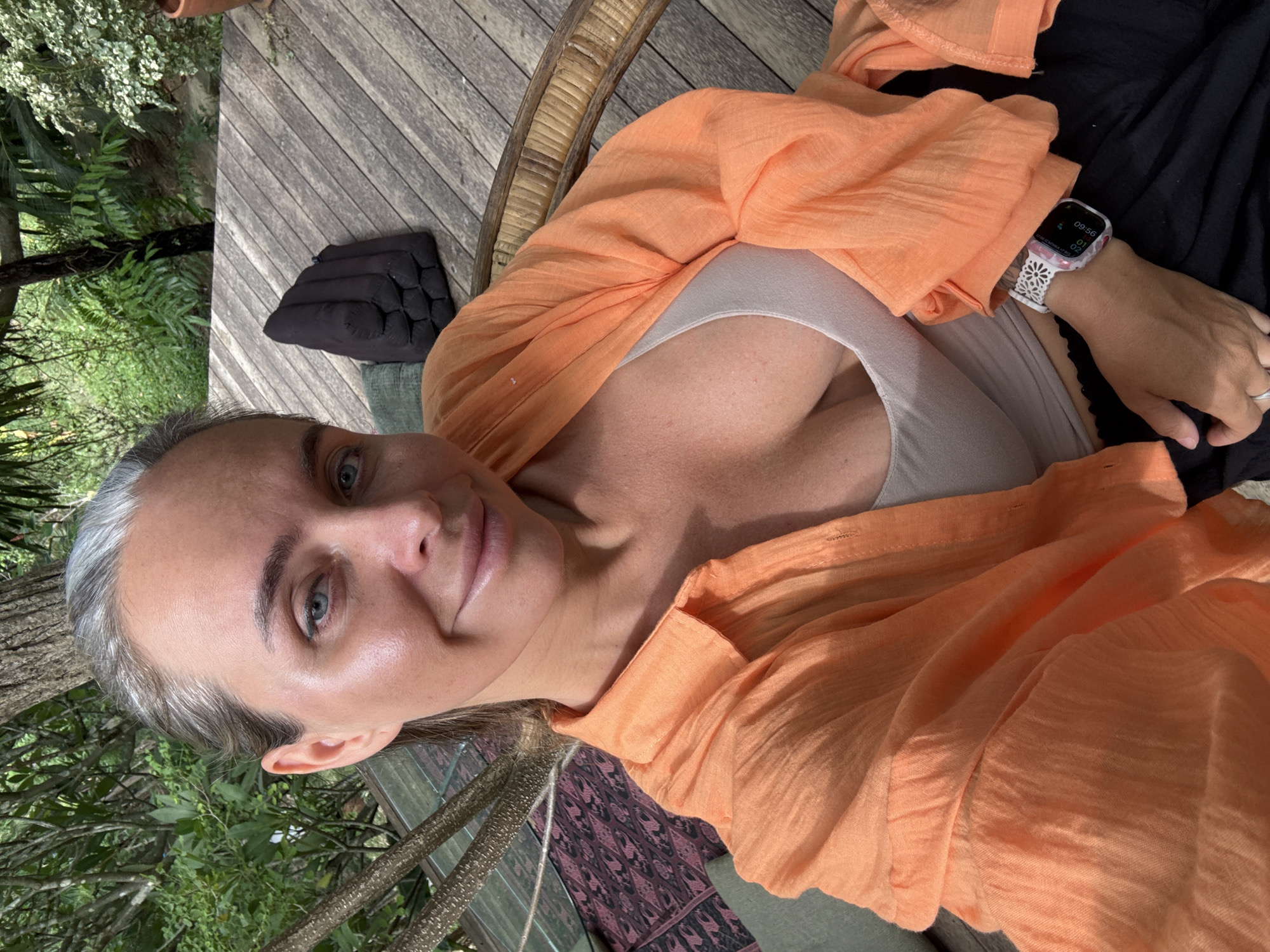A woman with graying hair and blue eyes takes a selfie outdoors. She is wearing a light-colored tank top and an open orange button-up shirt. Her left arm rests on her lap, displaying a smartwatch with a white band and pink floral pattern. She is seated on a wicker chair on a wooden deck. In the background, lush green foliage and trees suggest a tropical setting, consistent with Ko Samui District, Thailand. A black cushion and a patterned textile are visible on the deck.