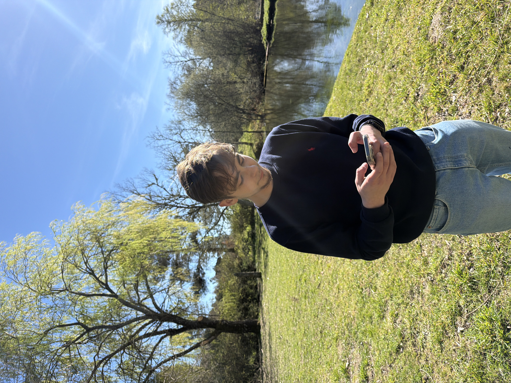 In an outdoor daytime scene in Vila Real, Portugal, a male individual stands on a grassy area beside a body of water. Positioned slightly left of center, he wears a navy blue long-sleeved sweatshirt with a small red polo logo on the left chest, and light blue denim jeans. He has light brown hair and subtle facial stubble. Both hands hold a silver-cased smartphone, upon which his gaze is directed downward, indicating interaction with the device. The background features a reflective body of water to the right, bordered by a variety of trees. Some trees display fresh green spring foliage, while others appear to be in an earlier stage of budding. The sky above is clear and bright blue with faint high clouds, and a distinct sun flare is visible in the upper right quadrant of the image.