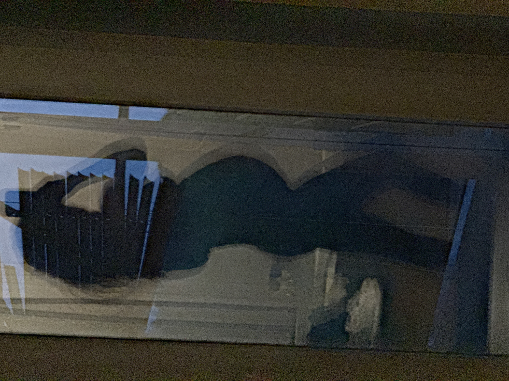 The image appears to be a reflection in a window, possibly a window display. The reflection shows the silhouette of a person standing or sitting in a chair. Behind the person, there's what looks like a radiator or a similar metal structure with vertical elements. The reflection is somewhat blurred and overlaid with the view through the window, which shows indistinct architectural details and a light-colored surface. It's difficult to ascertain specific details about the location or the individuals due to the reflection and low clarity. The image does not contain sufficient information to confirm the city of Chaikovsky, Russia, as the location.