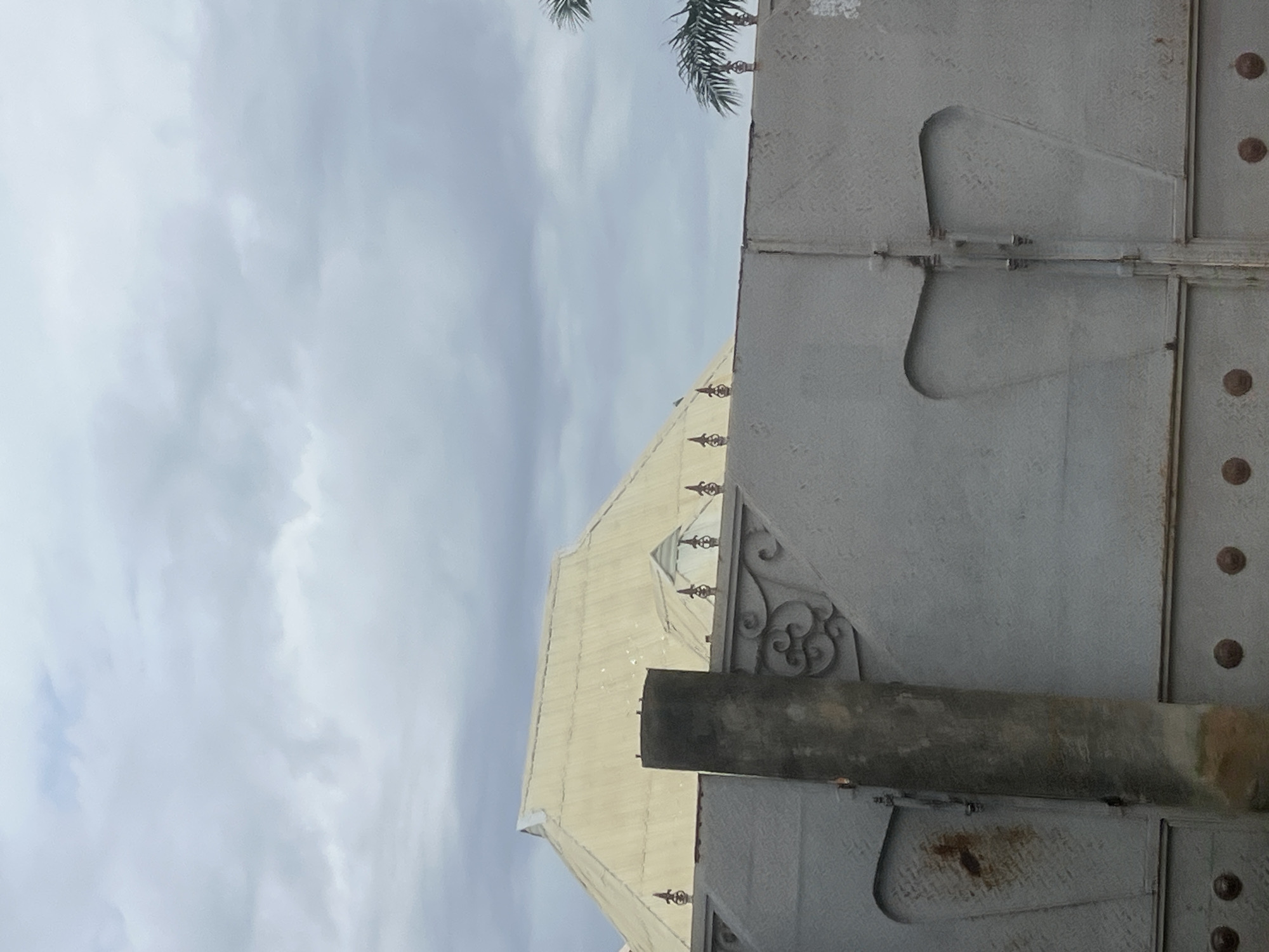 This image shows a grey metal gate with decorative elements. Behind the gate, a weathered, light-colored, multi-peaked roof structure of a building is visible. A portion of a concrete pillar is in the foreground, to the left of the gate. The sky is overcast with grey clouds. Palm fronds are visible on the far right edge of the image. This scene is located in Jalingo, Nigeria.