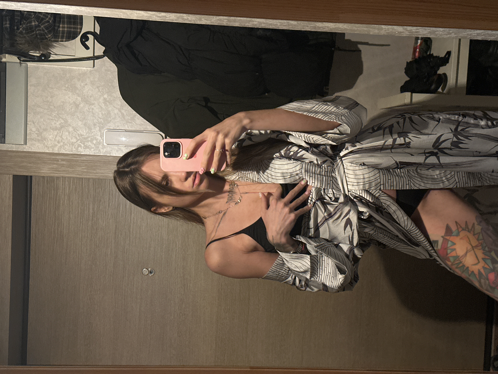 A young woman with long brown hair is taking a mirror selfie in a dimly lit room, possibly a hallway or dressing area, in Moscow, Russia. She is wearing a black camisole and a patterned robe that is open, revealing part of her torso and legs, which are tattooed. She is holding a pink smartphone in her right hand, angled to capture the reflection. The background includes a textured wall, a door, and a rack with clothing, including a dark puffer jacket and a plaid hat. A shelf with shoes and other items is also visible.
