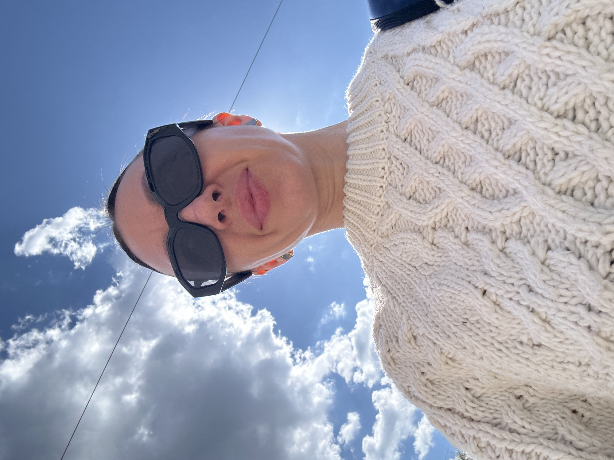 A woman is pictured from a low angle, wearing black sunglasses and a cream-colored cable-knit sweater. The background shows a sky with clouds, suggesting an outdoor setting. Power lines cut through the sky in the upper left. There is no indication in the image that the location is in Votkinsk, Russia.